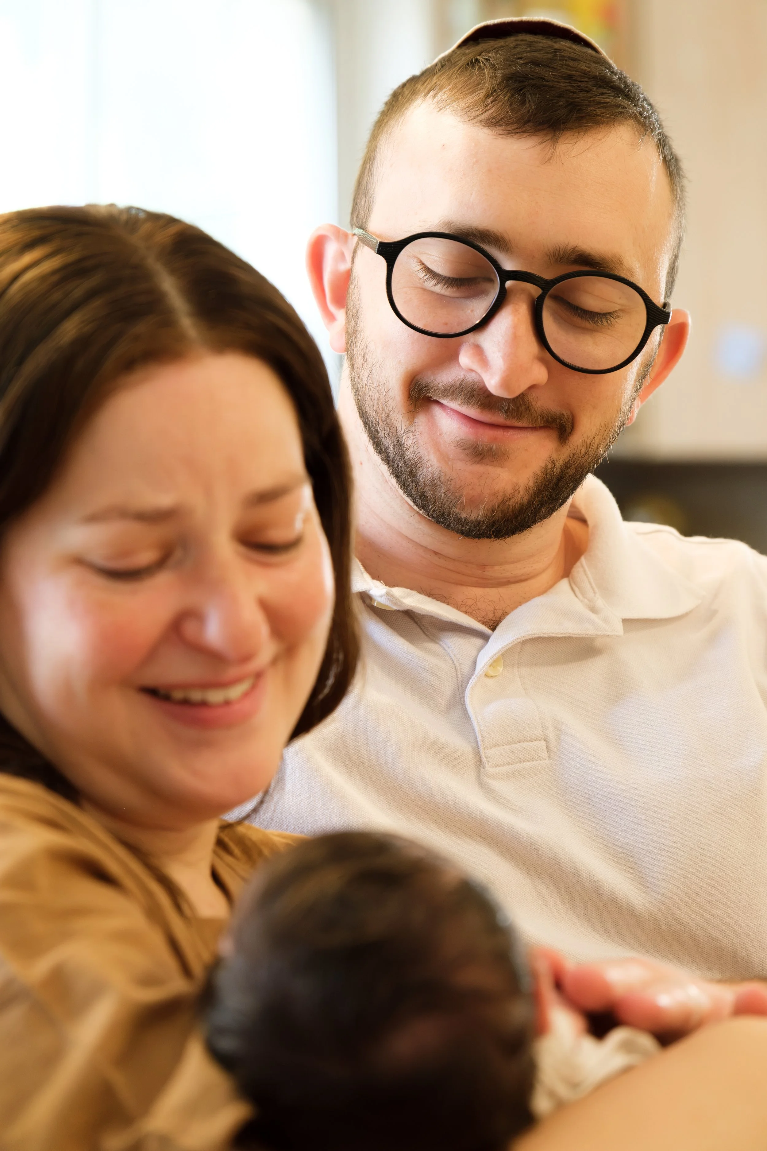 A smiling man with glasses and a beard, and a woman with brown hair, smiling and looking down, with children in the foreground, in an indoor setting.