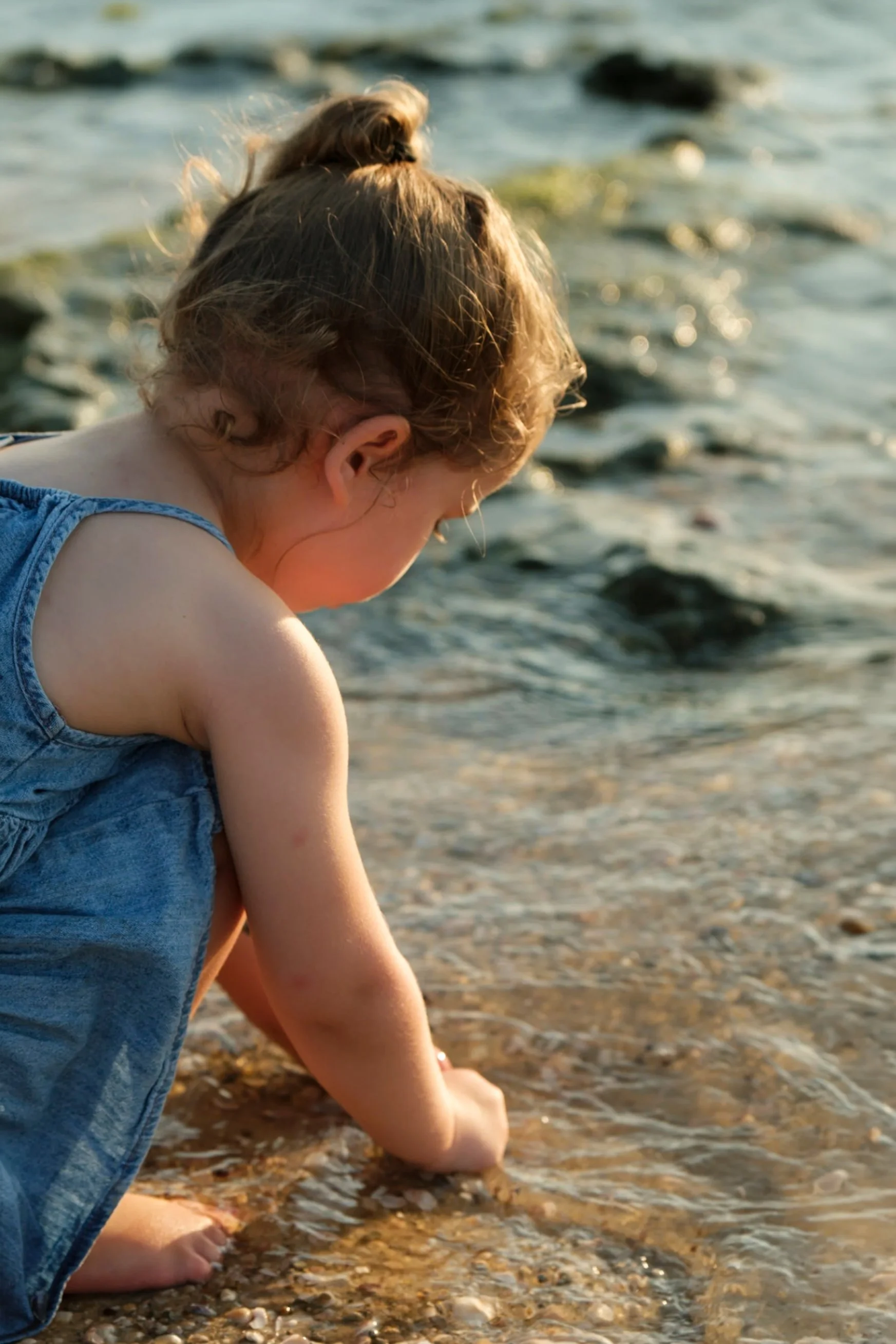 A young girl with curly hair and a blue sleeveless top sitting by a body of water, possibly a beach or lakeside, with rocks and pebbles on the shore during sunset.