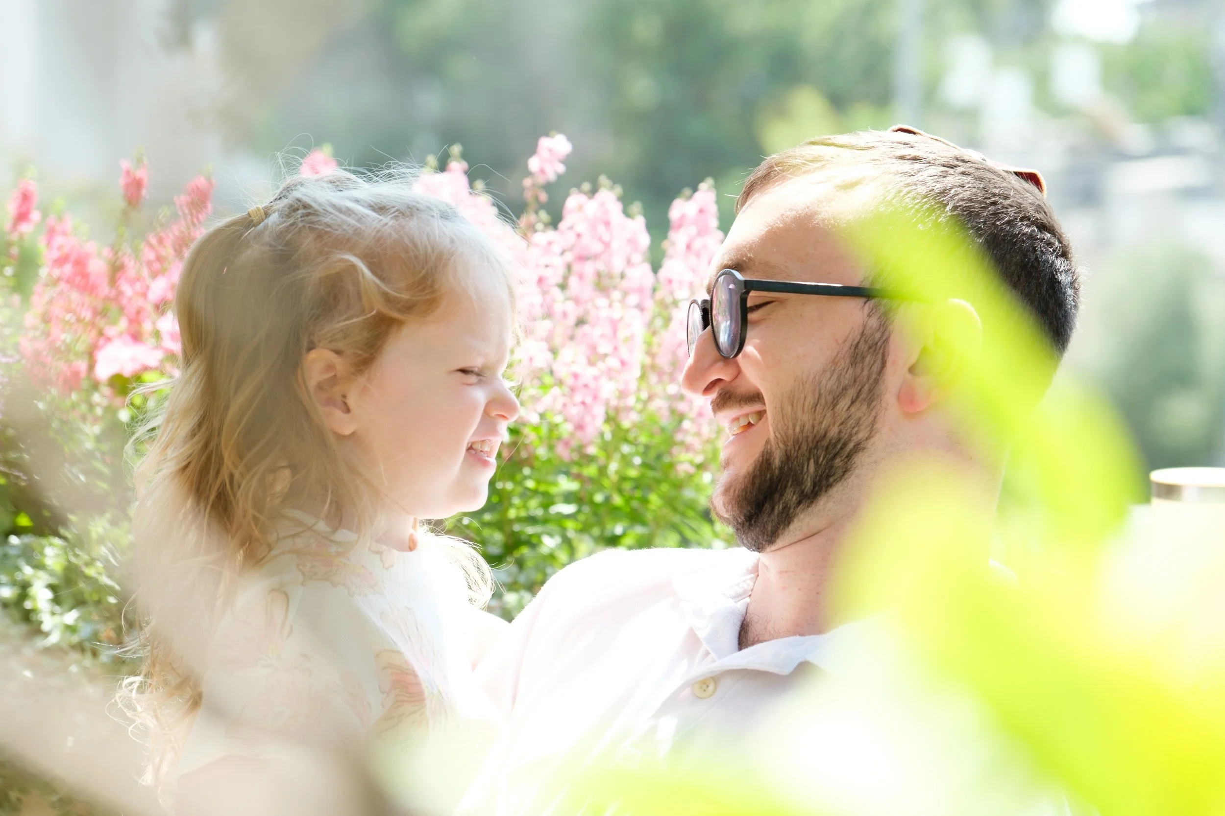 A man and a young girl smiling at each other outdoors with pink flowers and green foliage in the background.
