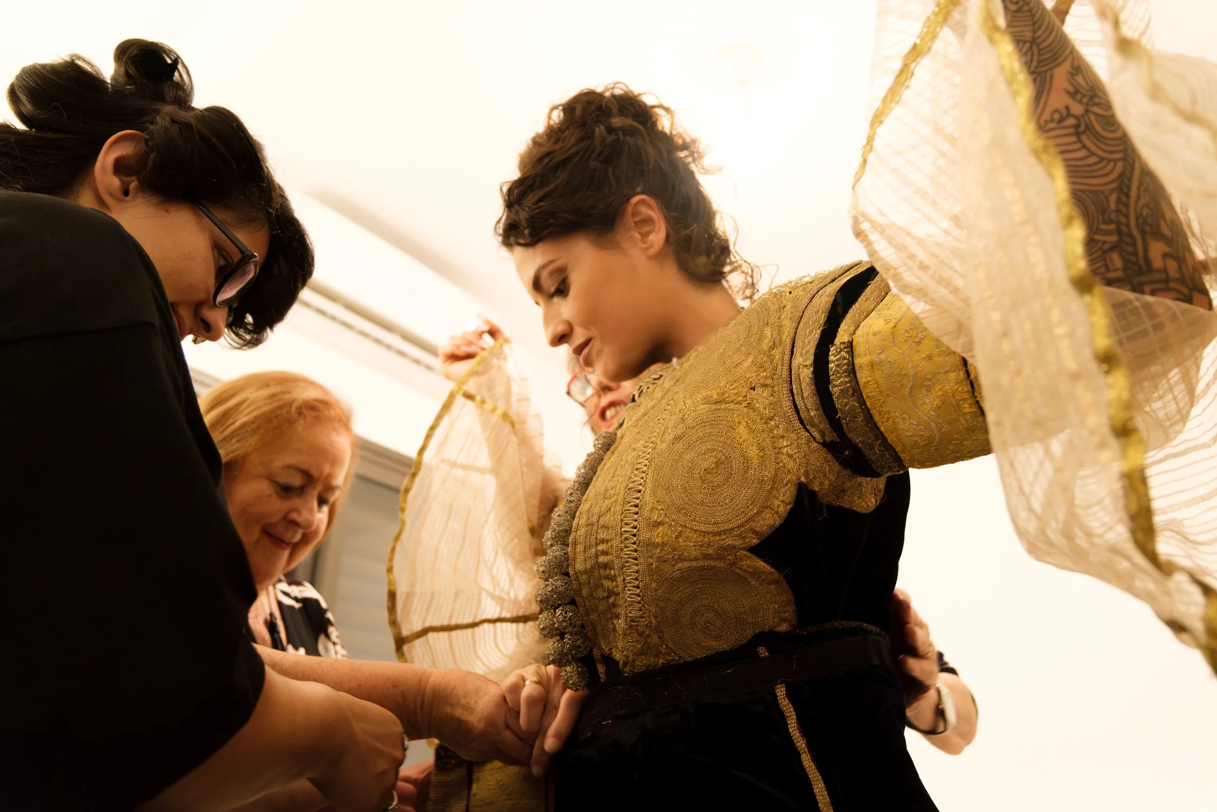 A group of women participating in a cultural or traditional ceremony, with one woman dressed in ornate gold fabric and others helping her with a task.