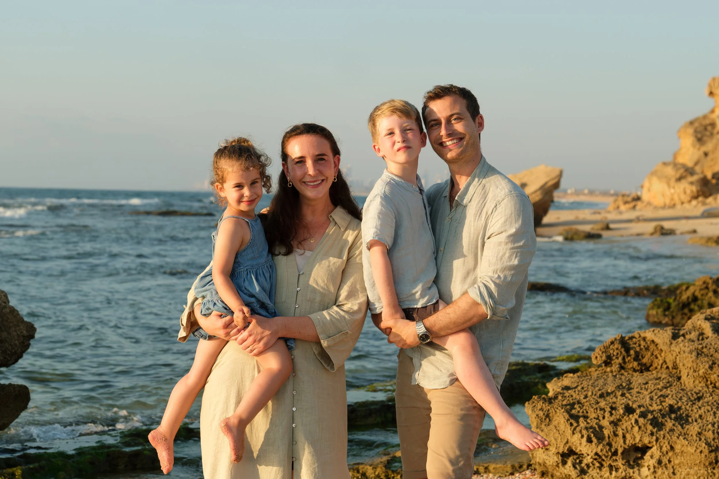Family of four enjoying a day at the beach with rocks, ocean waves, and a clear sky in the background.