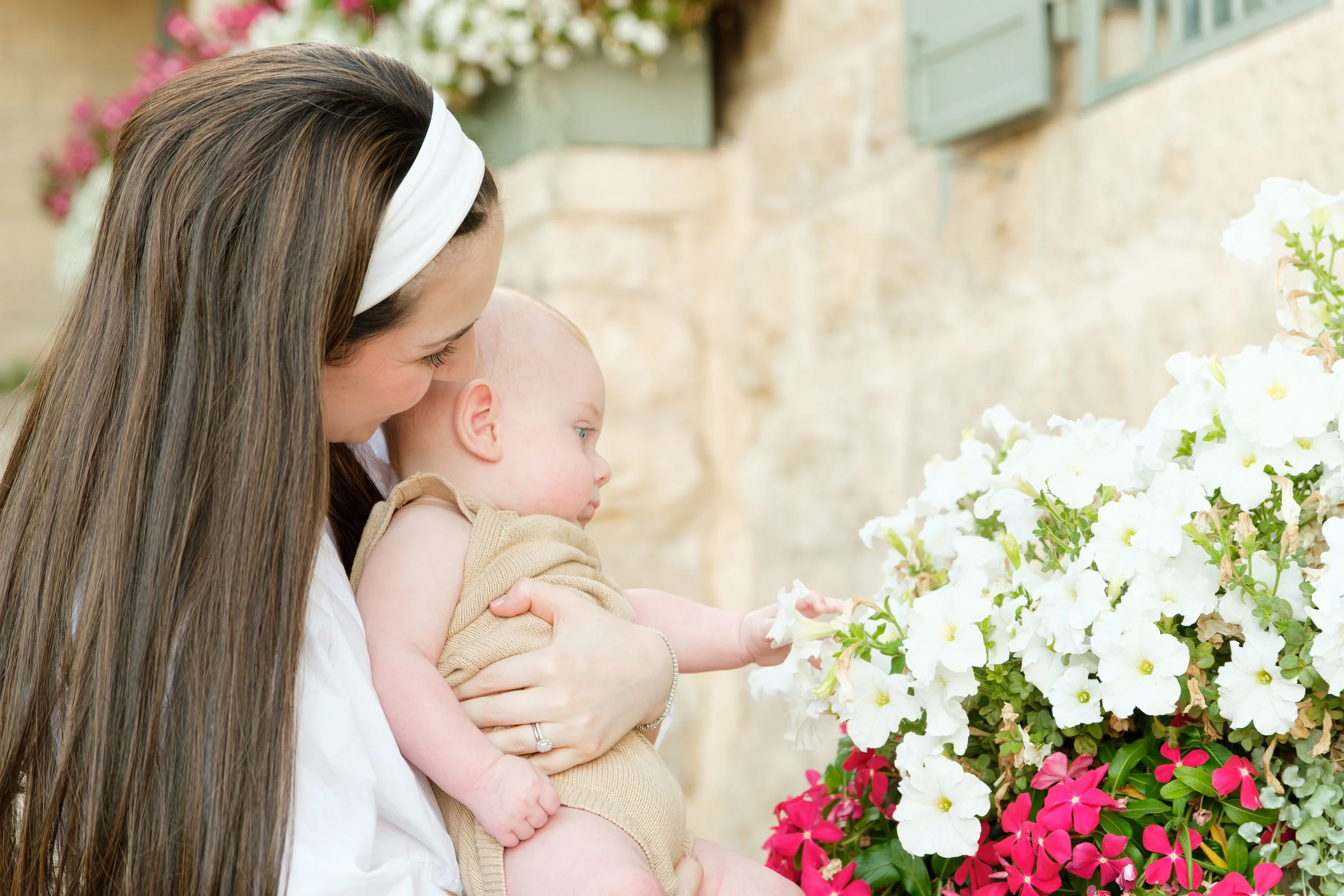 A woman with long brown hair wearing a white headband holding a young child, both looking at a flower arrangement of white, pink, and red flowers outside on a sunny day.