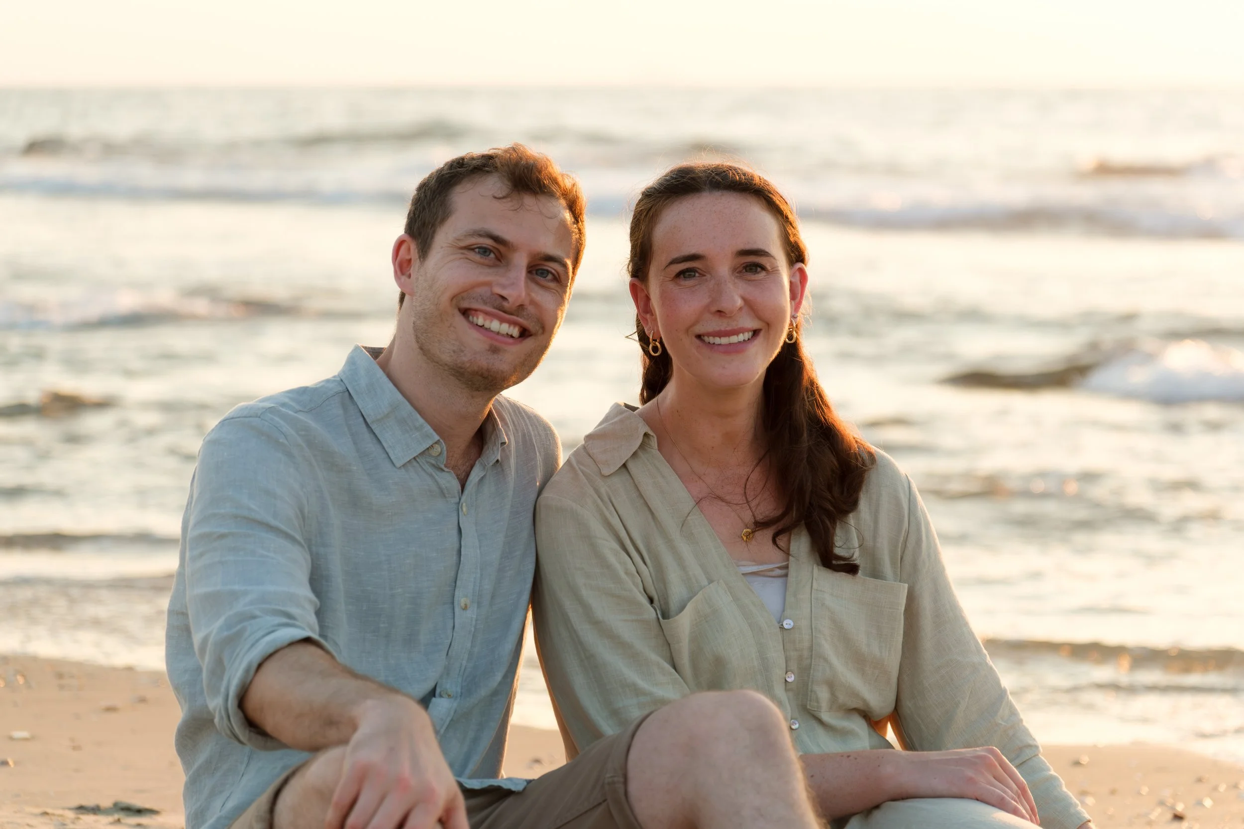 A smiling couple sitting on the beach at sunset, near the ocean, with waves in the background.