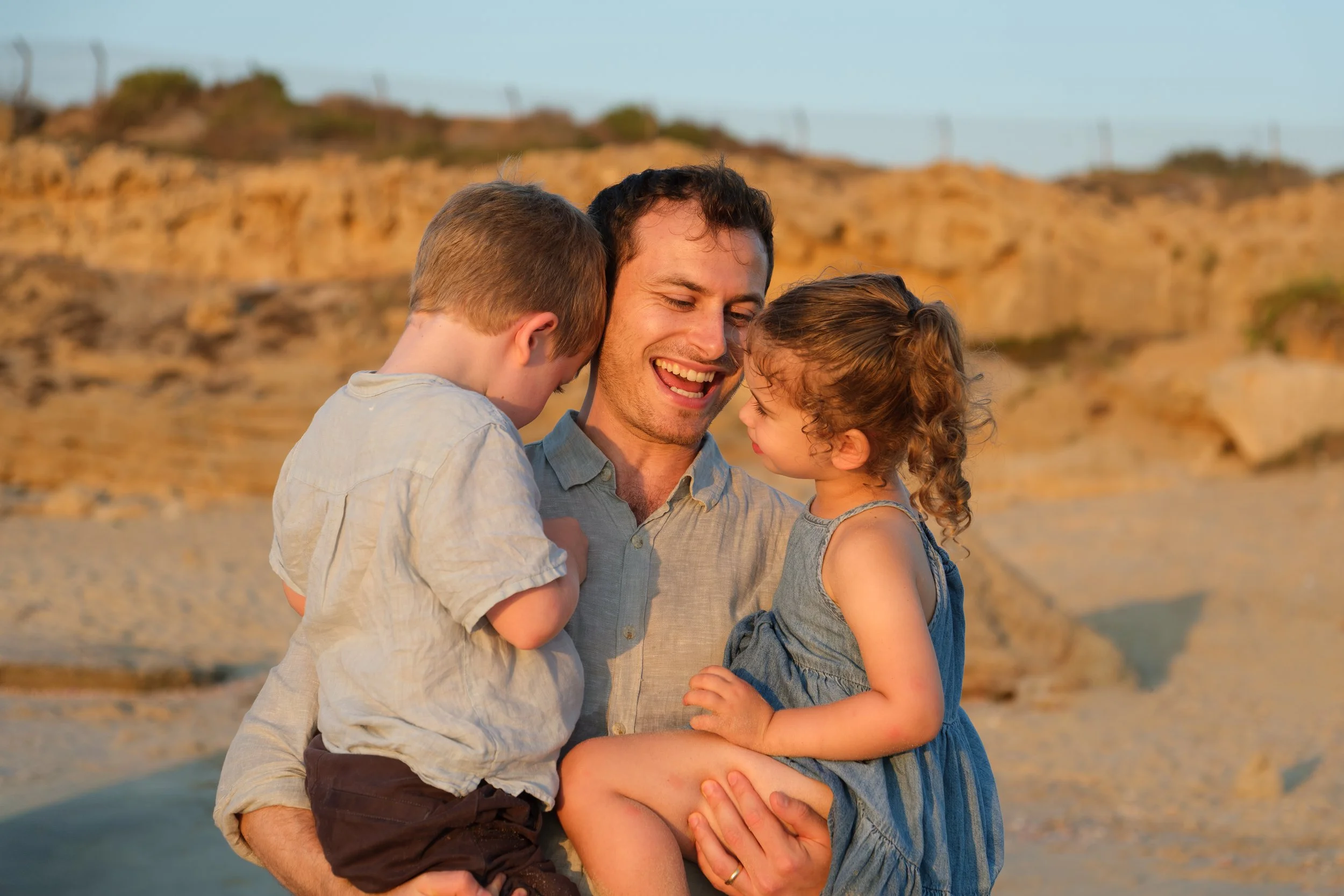 A man is holding two children, a boy and a girl, they are hugging and smiling at each other on a beach at sunset with rocky cliffs in the background.