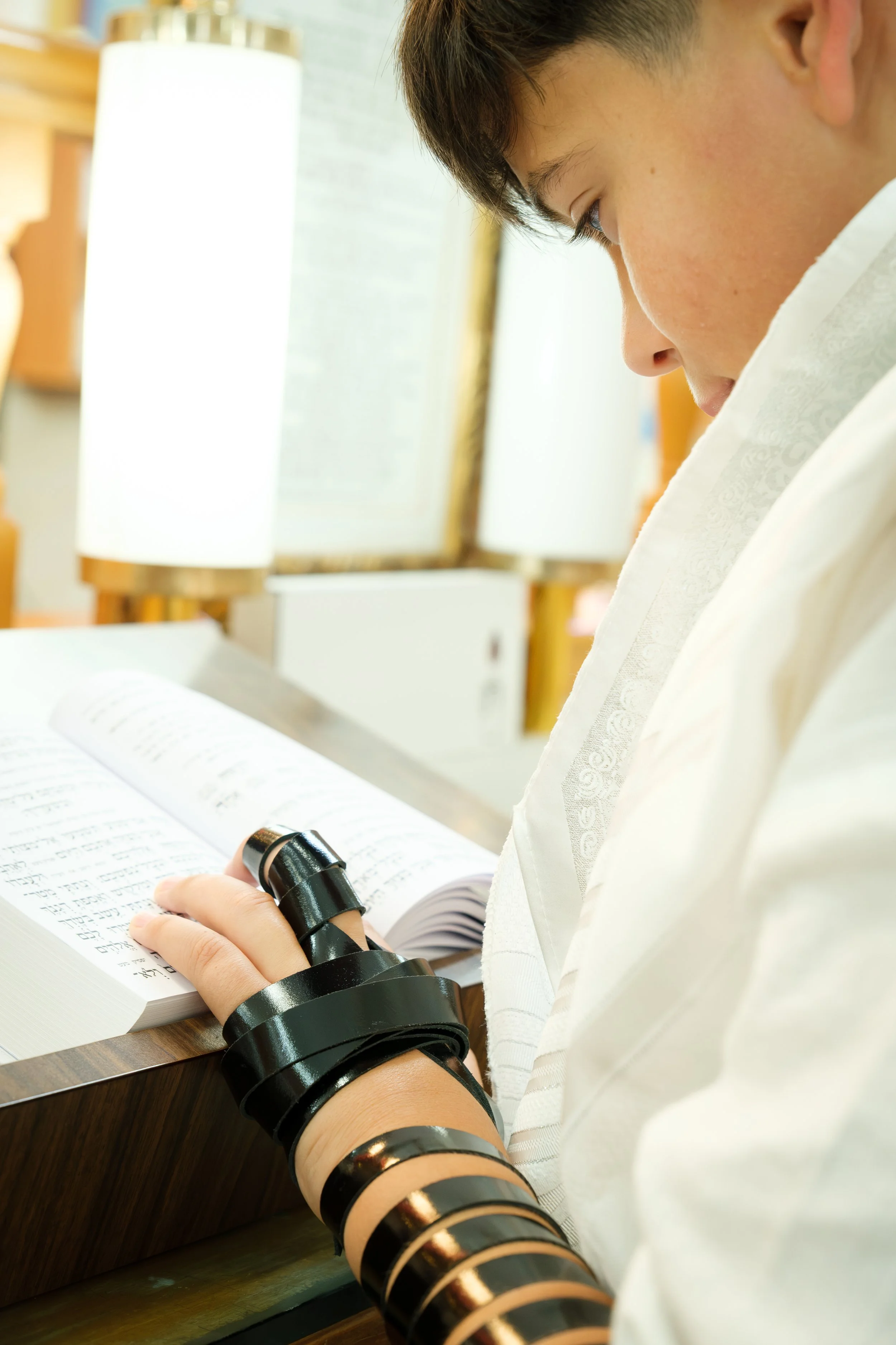 A person with dark hair and a white garment is reading a book with text in a language that appears to be Hebrew. The person is wearing multiple black bangles or cuffs on their left arm and is holding a pair of black glasses.