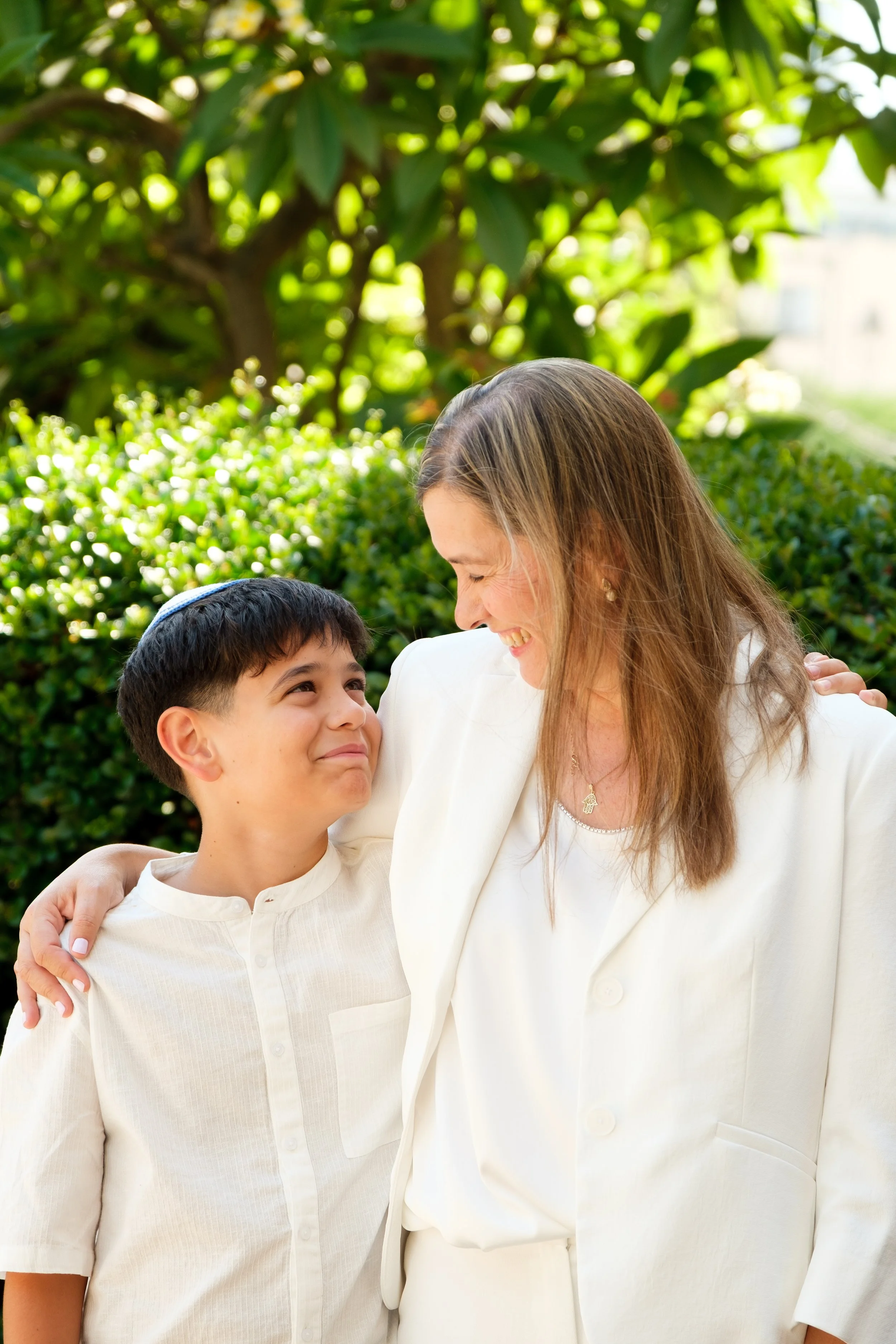 A woman and a boy smiling at each other outdoors, with green shrubs and trees in the background.