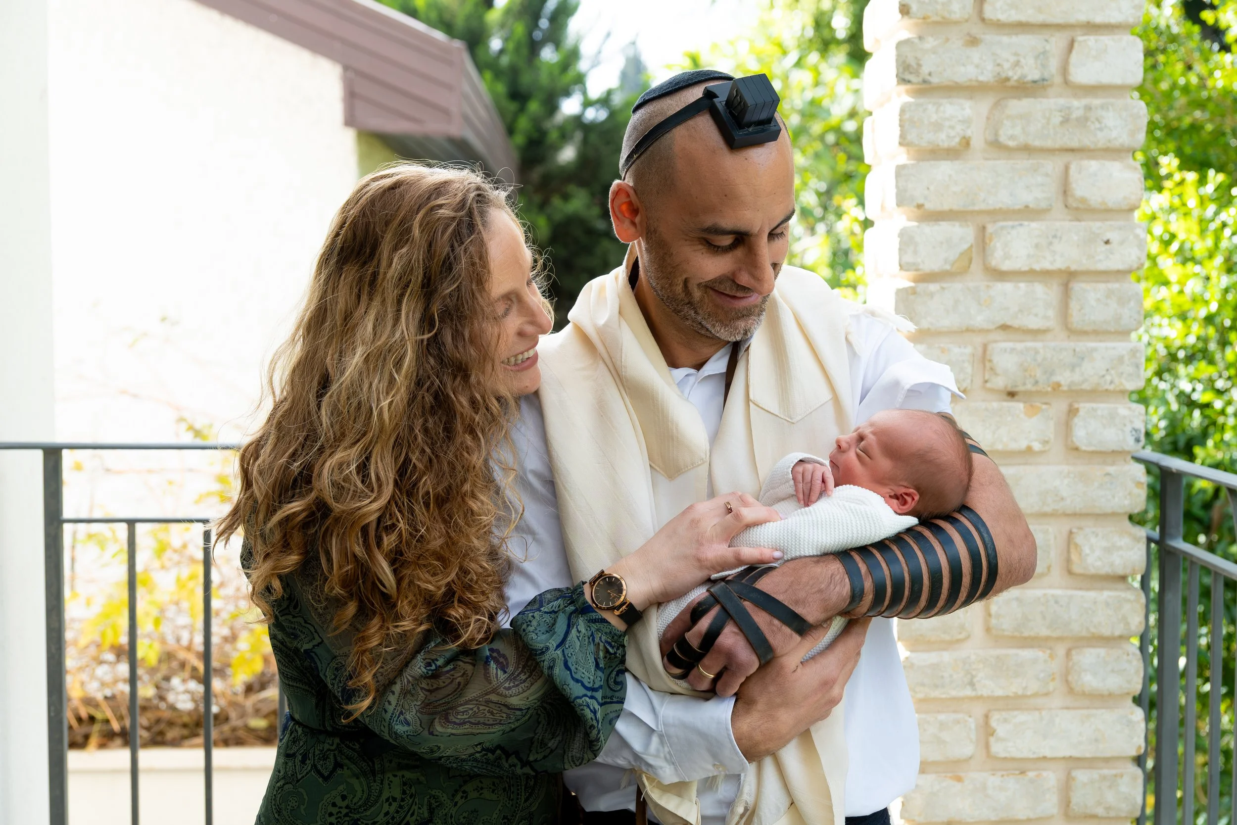A man holding a swaddled newborn baby while woman with long curly hair looks on smiling in an outdoor setting with trees and a brick pillar.
