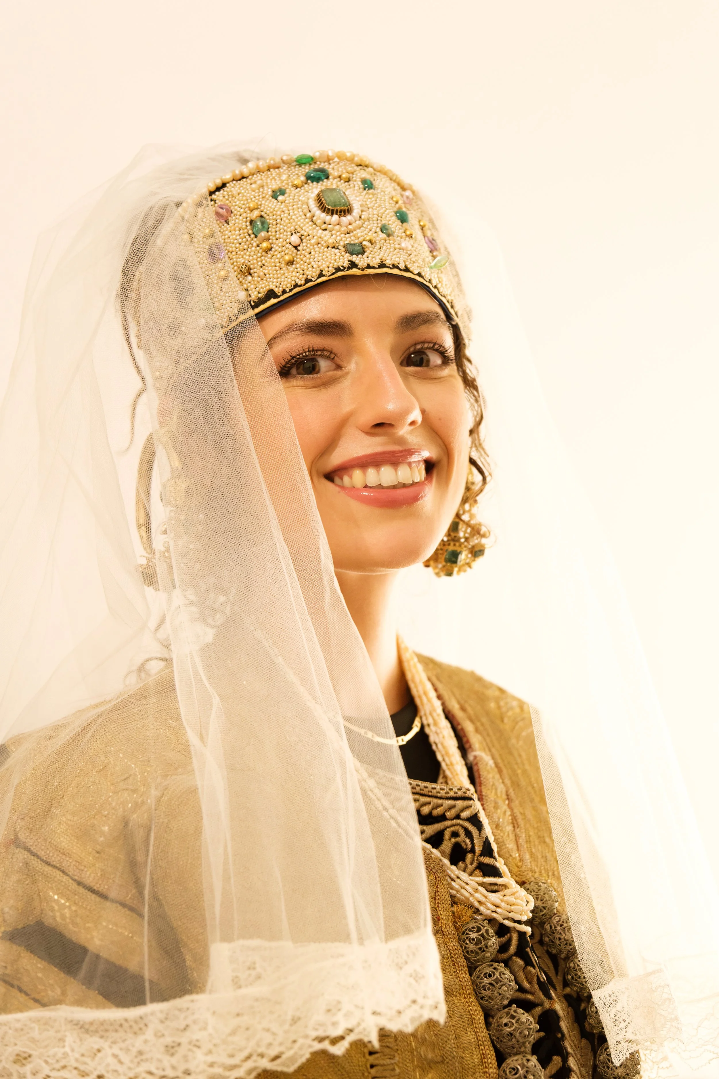 A woman wearing a beaded headdress with pearls and gemstones, veiled with lace, smiling and dressed in a traditional, ornate outfit.