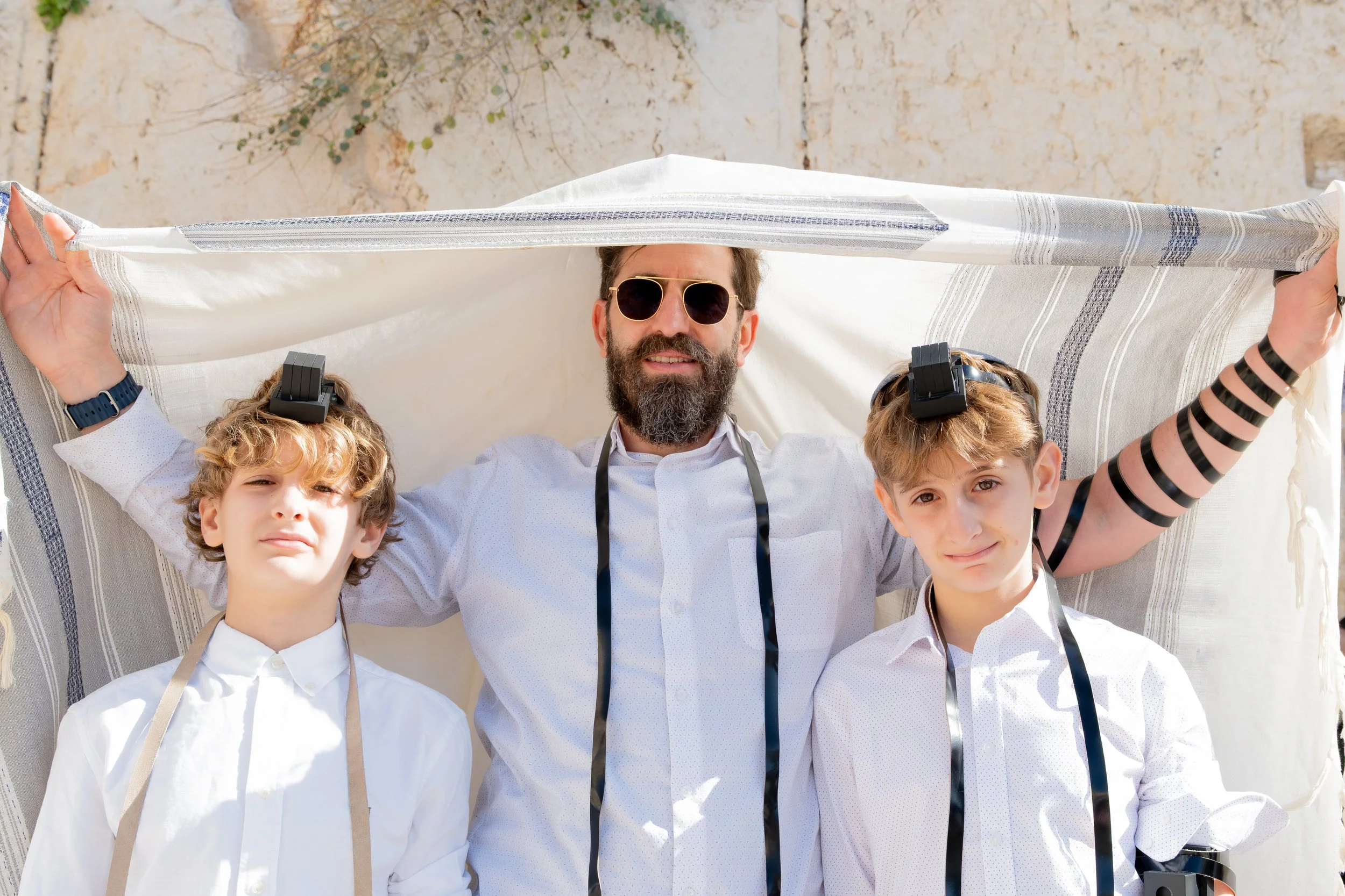 A man with a beard and sunglasses looking up, holding a fabric roof over three young boys dressed in white shirts, standing outdoors in front of a stone wall.