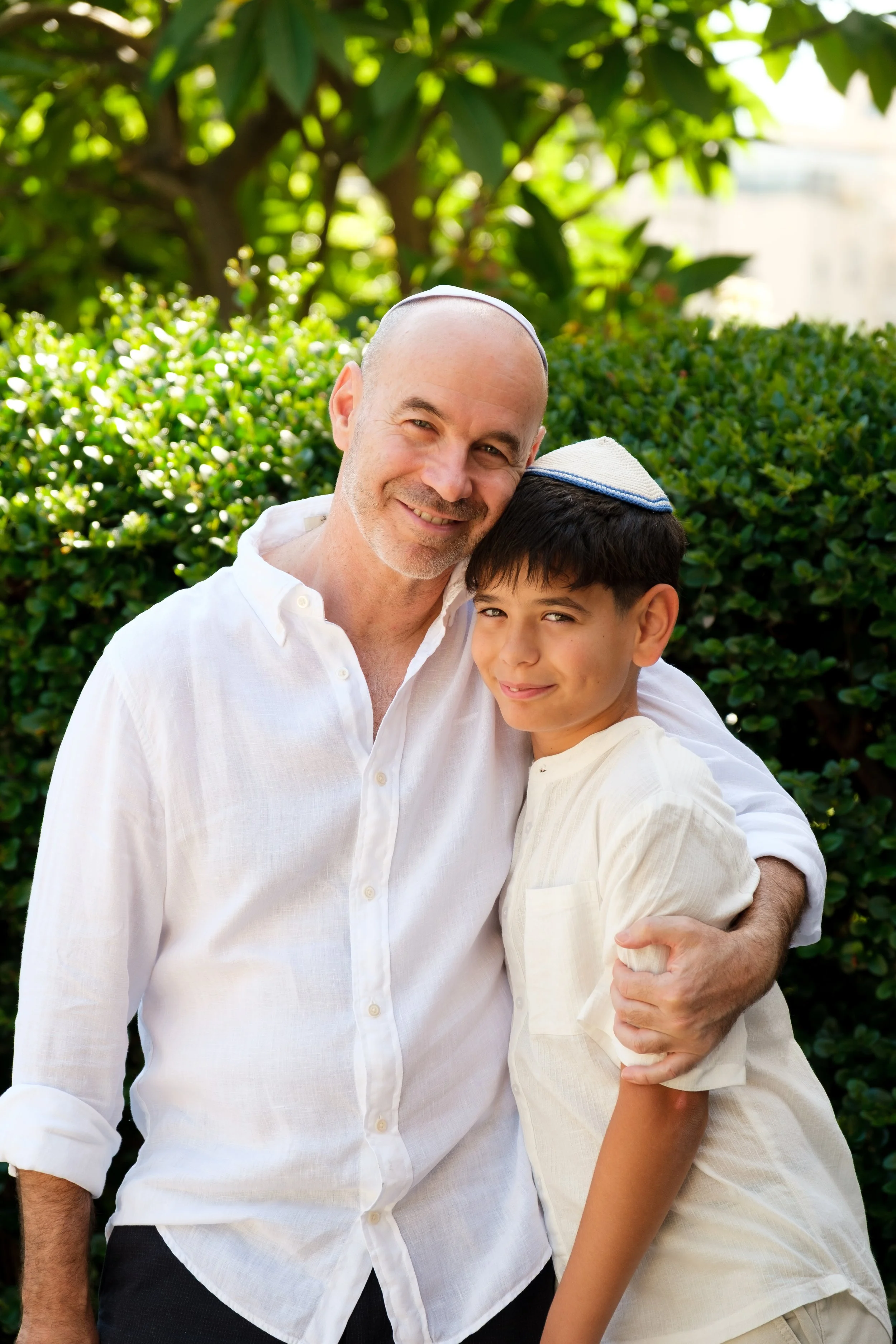 A man and a boy wearing kippahs, smiling and hugging outdoors with greenery in the background.