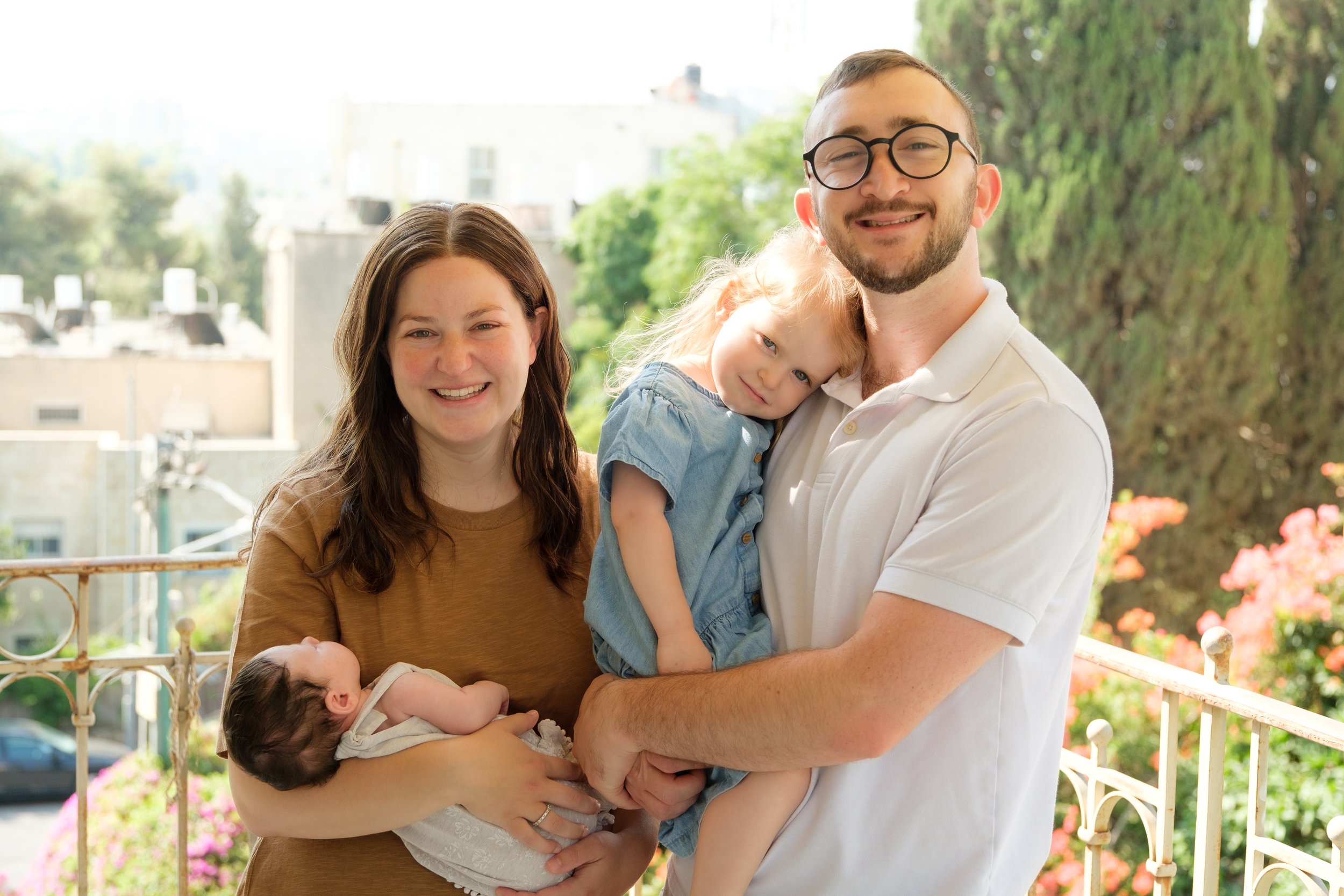 Family outdoors on a balcony with urban background, woman holding a newborn, man with glasses holding a toddler.