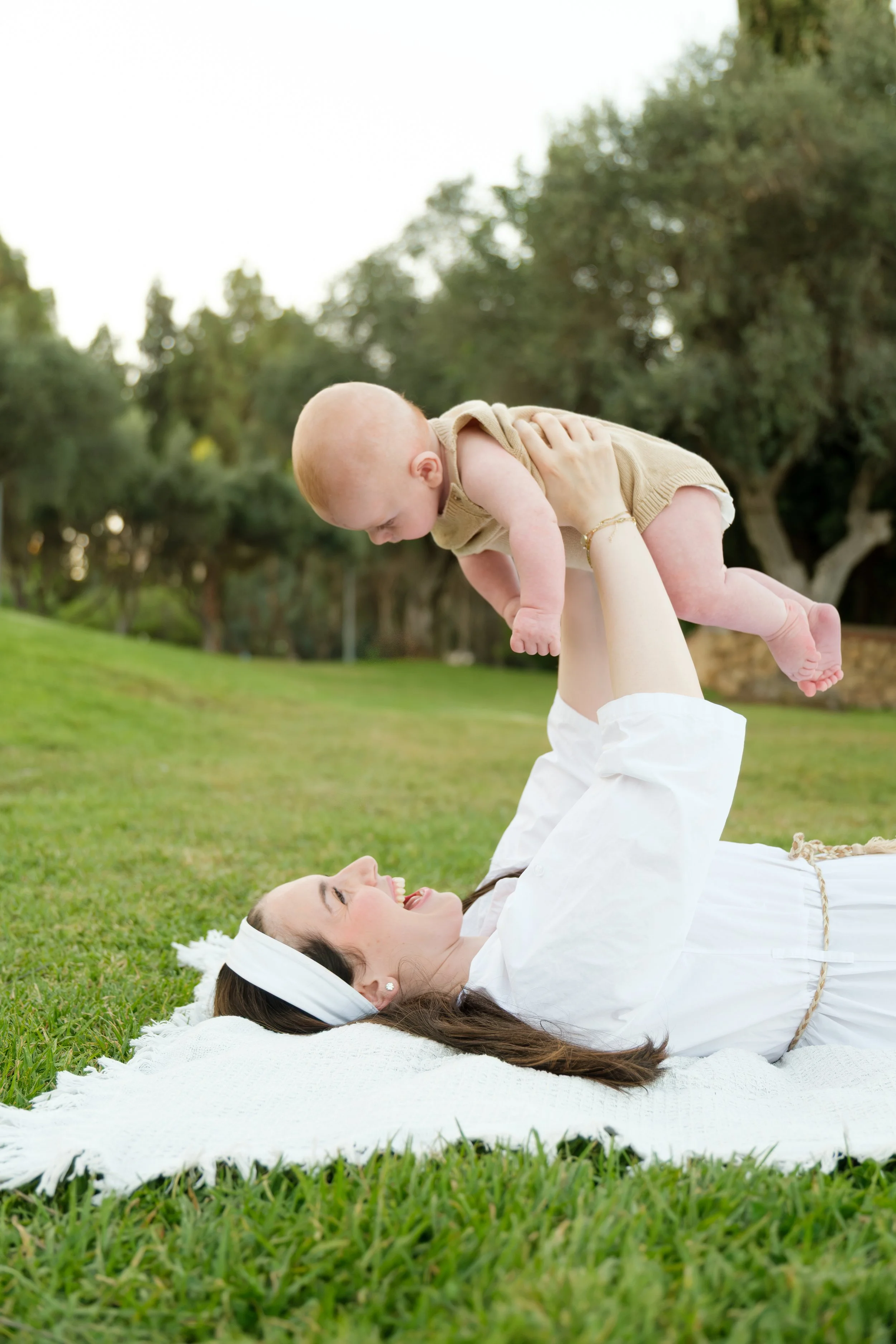 A woman lying on her back on a white blanket in a grassy park, playing with a baby boy who is being held up in the air by her hands, both smiling and looking at each other with trees in the background.