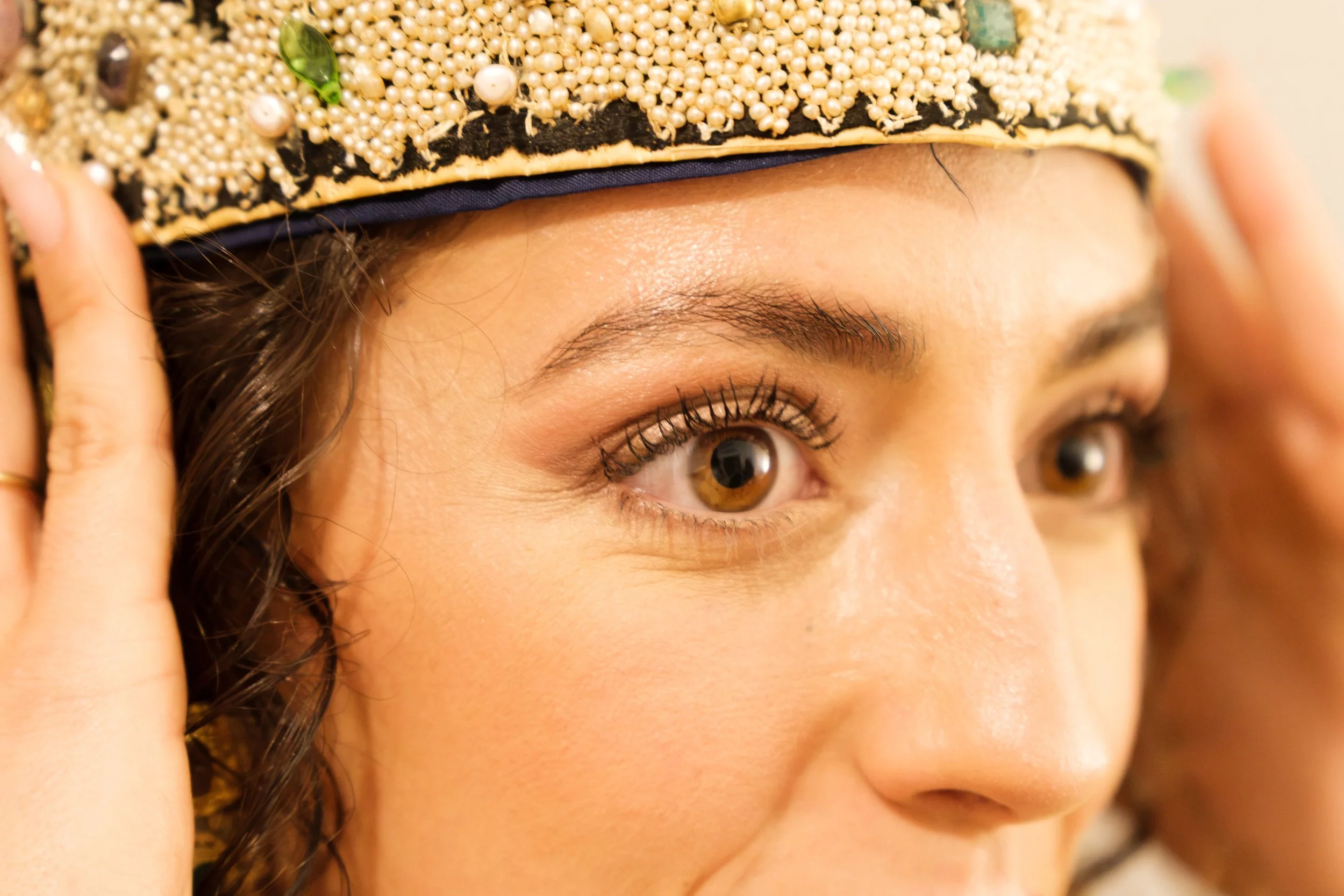 A person wearing a decorative hat embellished with beads, pearls, and green stones, touching the side of the hat with their fingers, with a close-up view of their face.