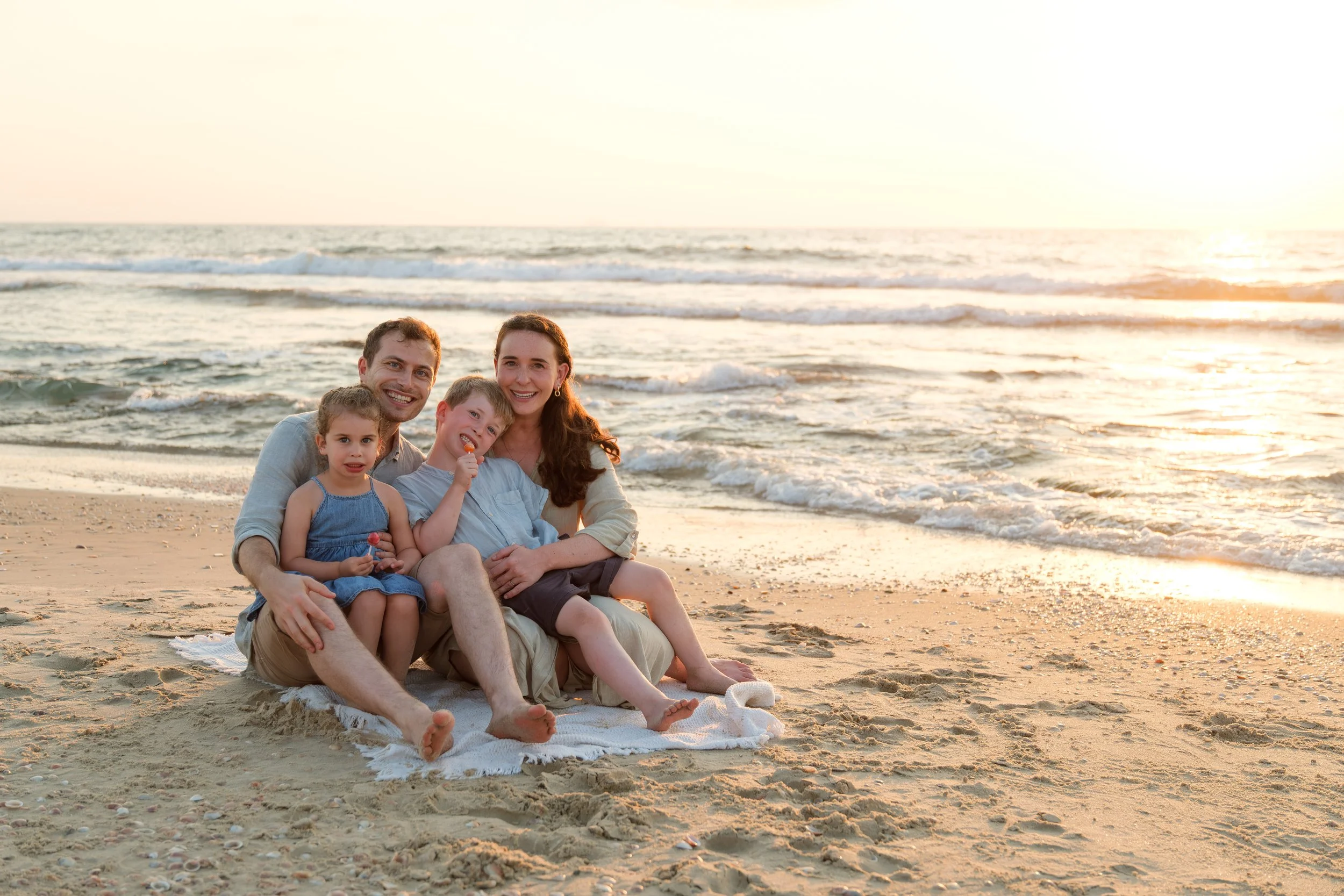 Family of four sitting on a beach at sunset, smiling, with the ocean and waves in the background.