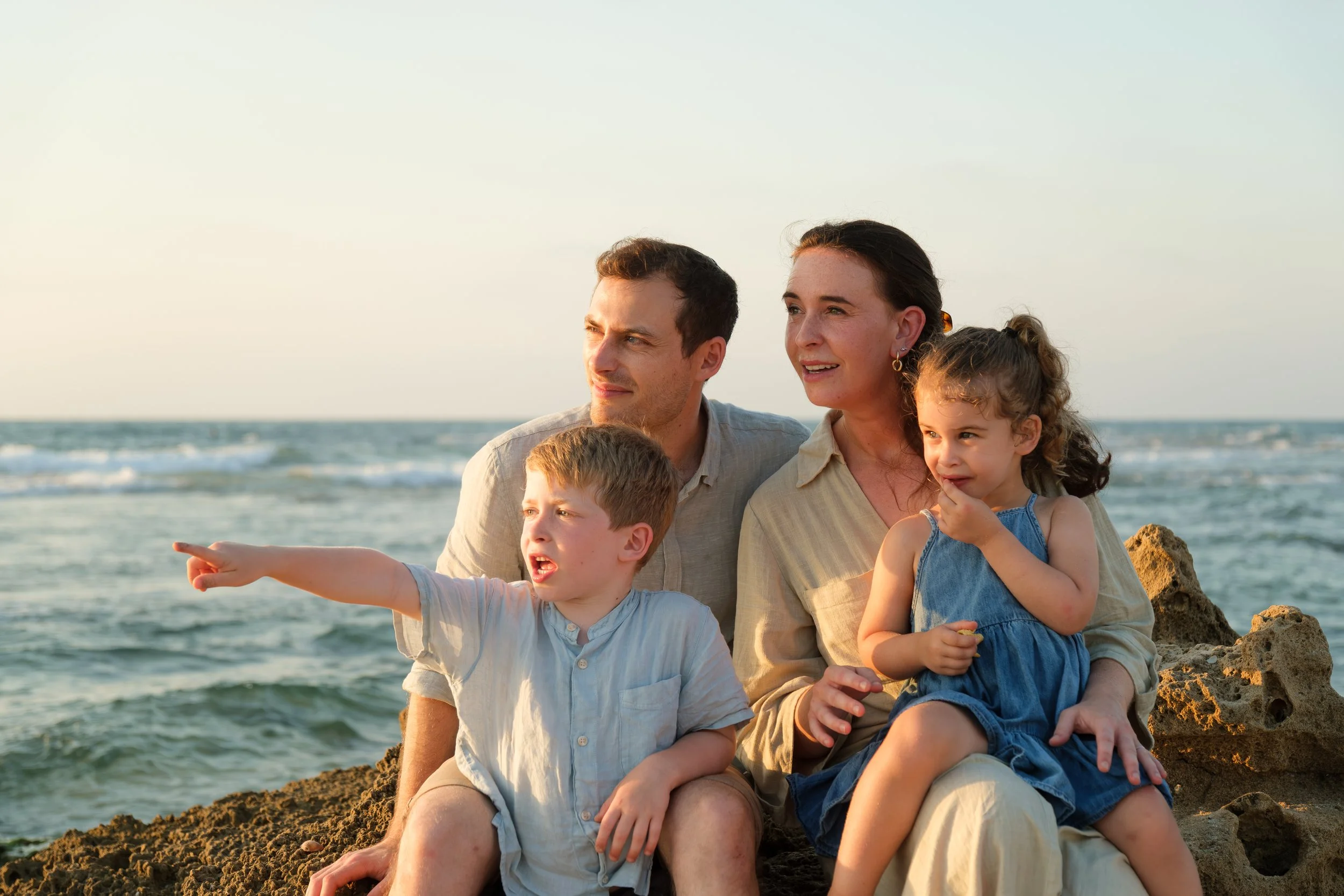 A family of four sitting on rocks by the beach during sunset, with the ocean and sky in the background. The adults and children are looking in different directions, with the child in the front pointing towards something in the distance.