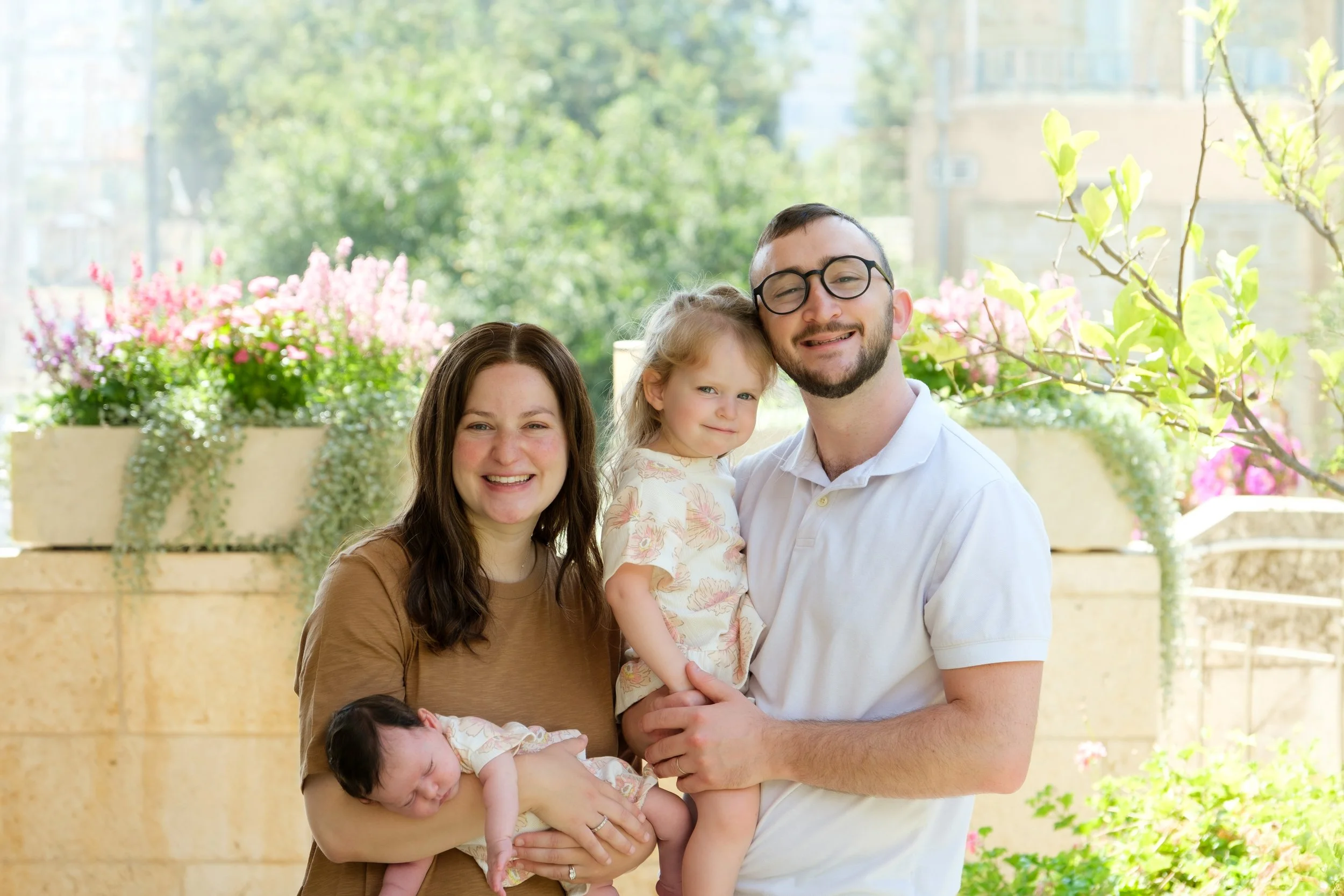 A happy family of four outdoors on a sunny day. The mother and father are holding their two young children, with pink flowers and green trees in the background.