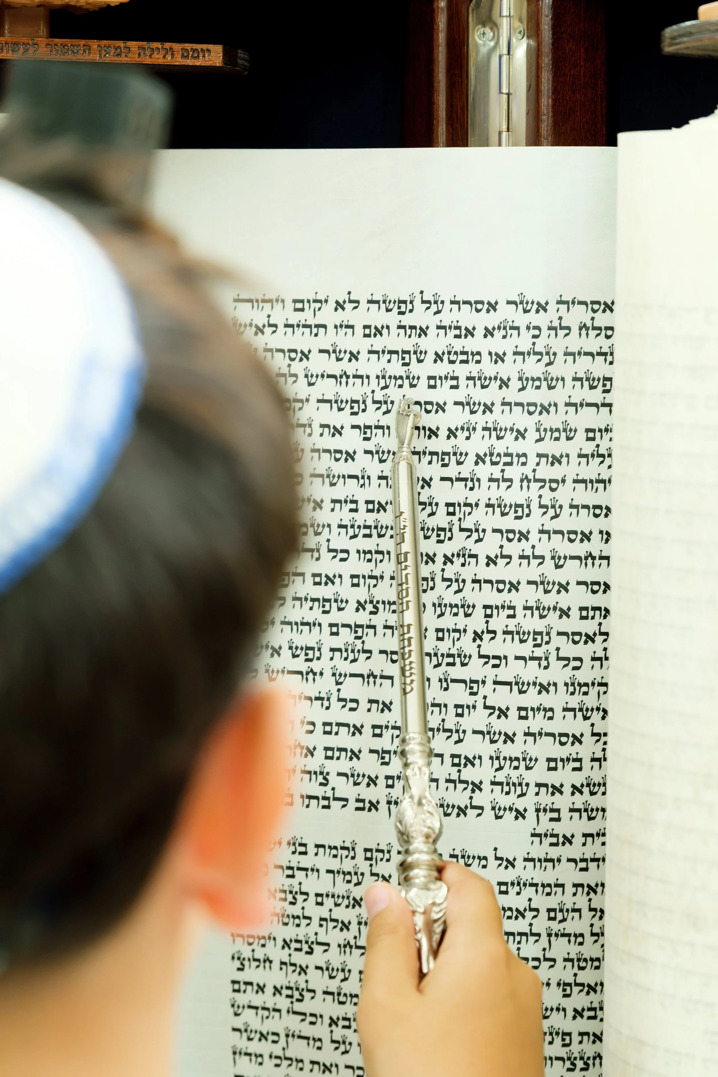 A person holding a silver prayer menorah in front of a Hebrew prayer book with Hebrew text, with a blurred child in the foreground.