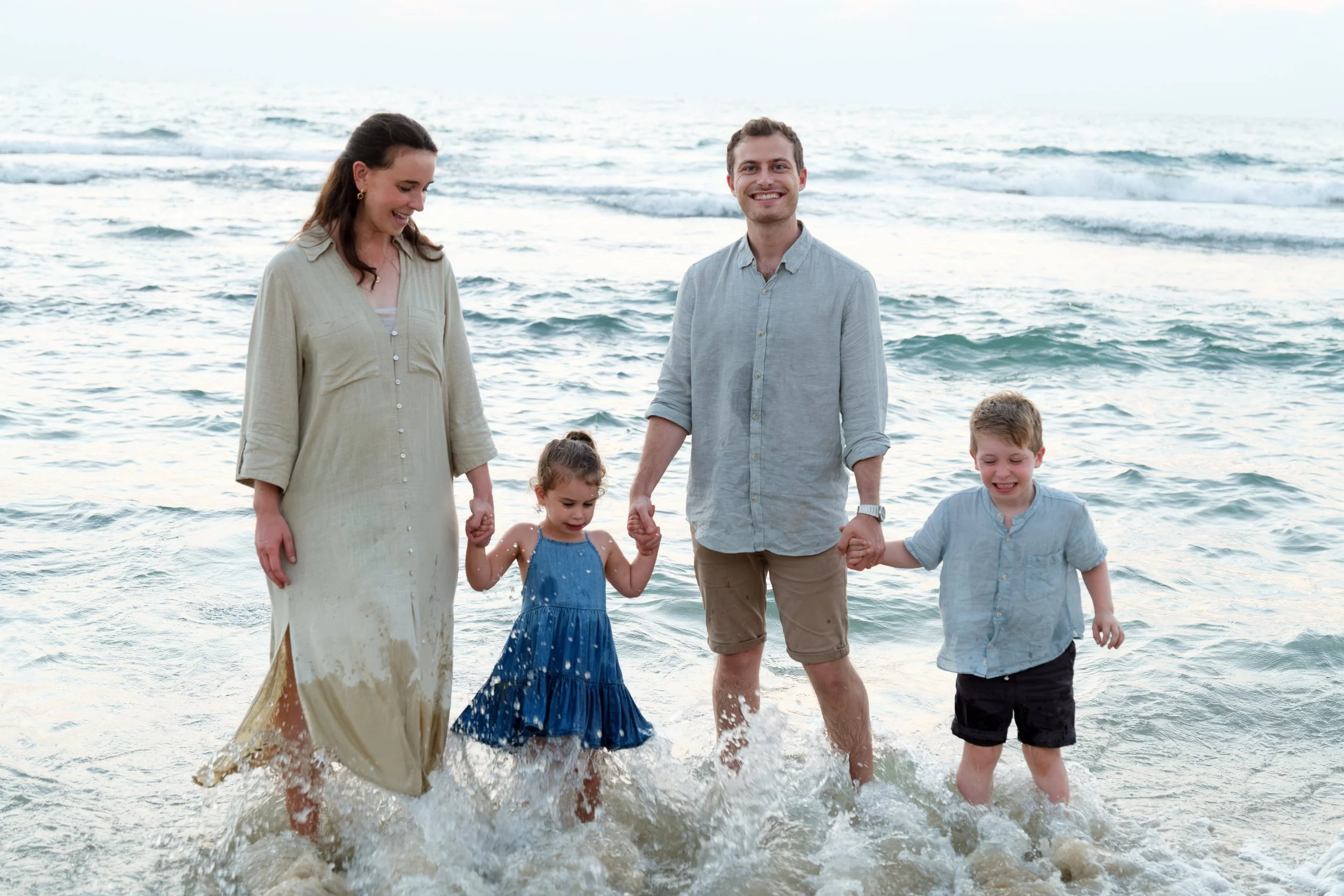 A family of five standing in the ocean at the beach. The mother, father, and three children are holding hands and smiling, with waves gently crashing around their feet.