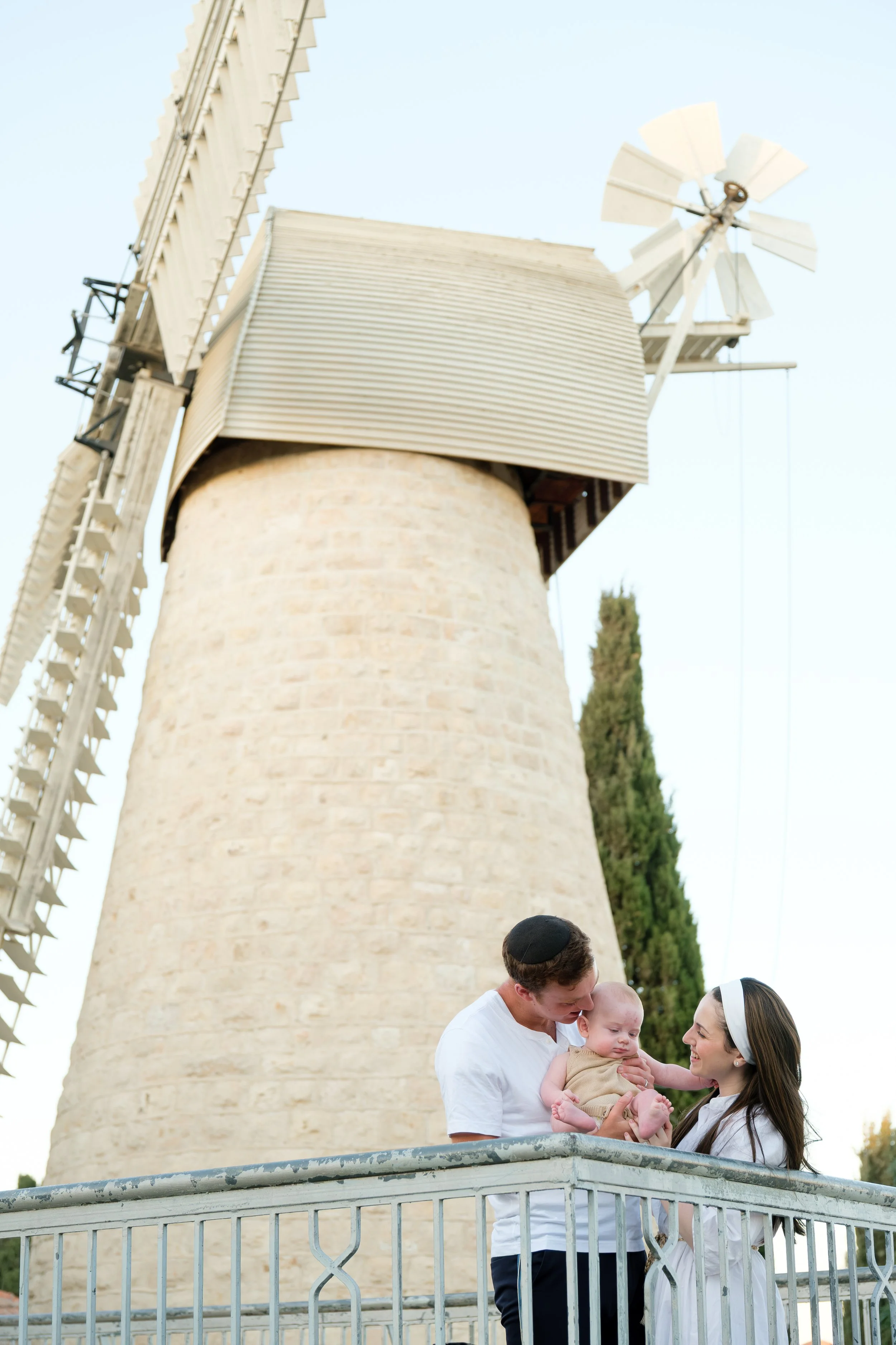 A Jewish family celebrating a religious event by a historic windmill. The man and woman, both wearing kippahs, are holding a baby between them on a platform with a decorative railing, smiling and looking at each other and the child. The windmill is m