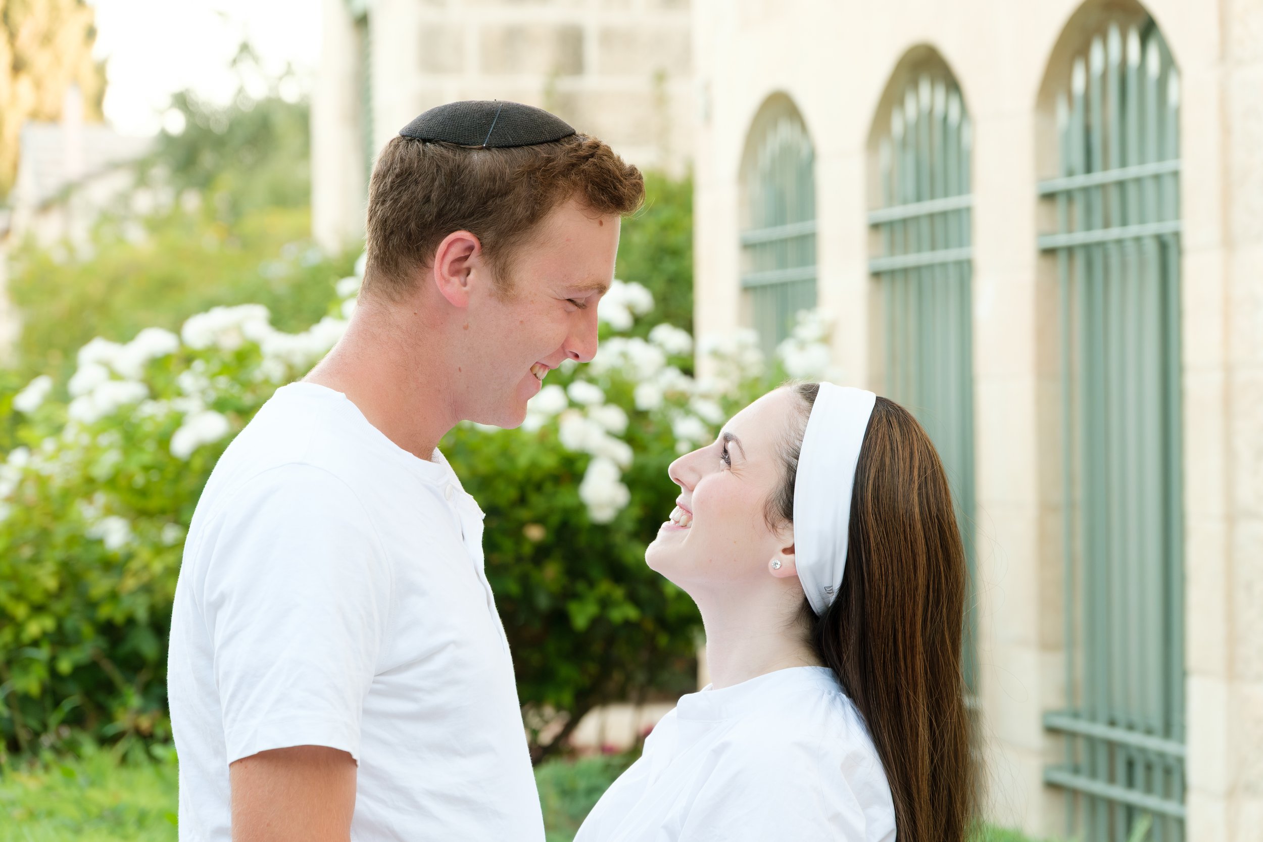 A young Jewish man and woman smiling at each other outdoors, with the man wearing a kippah and the woman wearing a white headband, in front of a building with arched windows and greenery.
