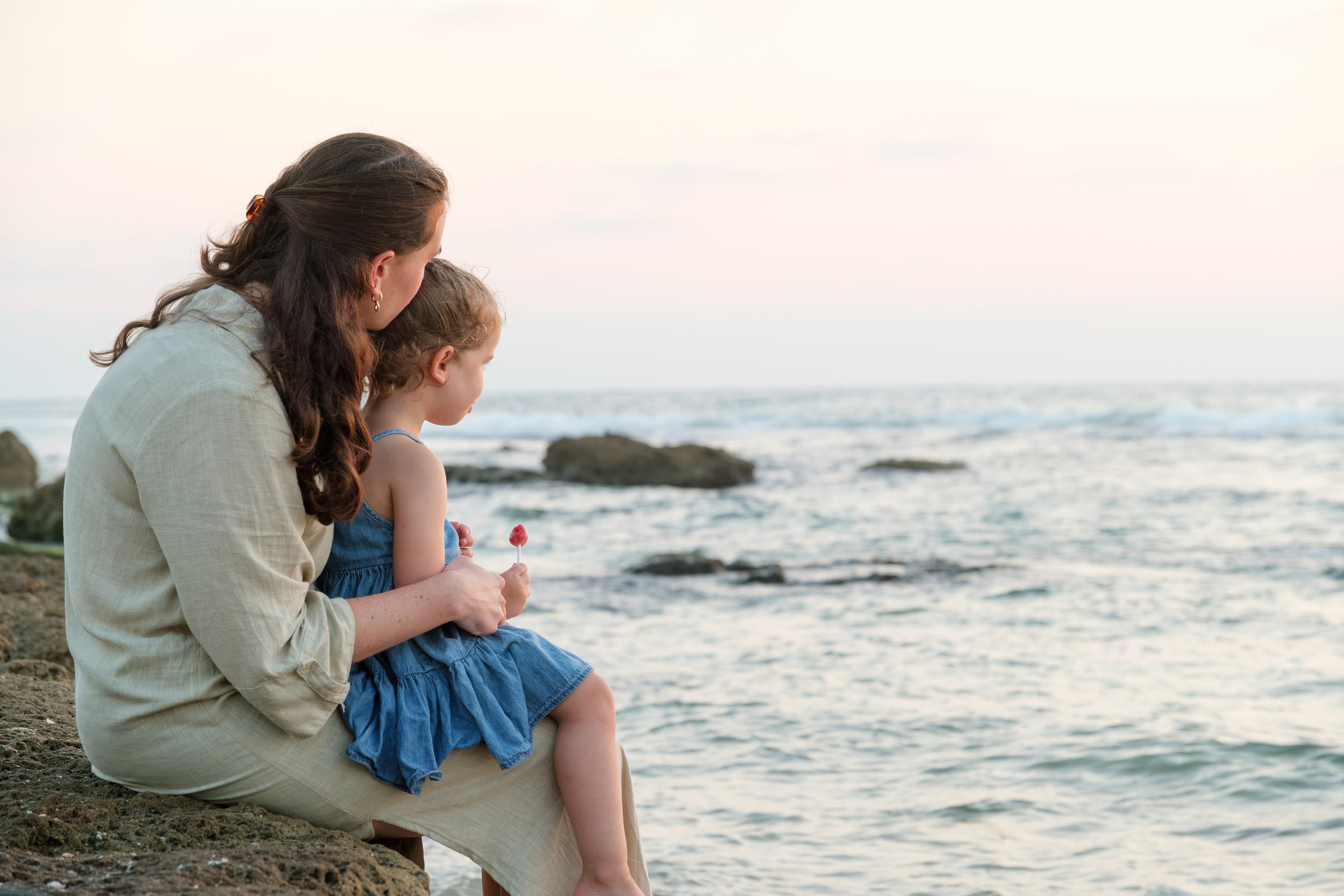 A woman and young girl sit on a beach at the shore, looking out at the ocean. The woman has long dark hair and is wearing a light-colored shirt, while the girl has curly hair and is wearing a blue dress, holding a pink lollipop. Rocks and the ocean w