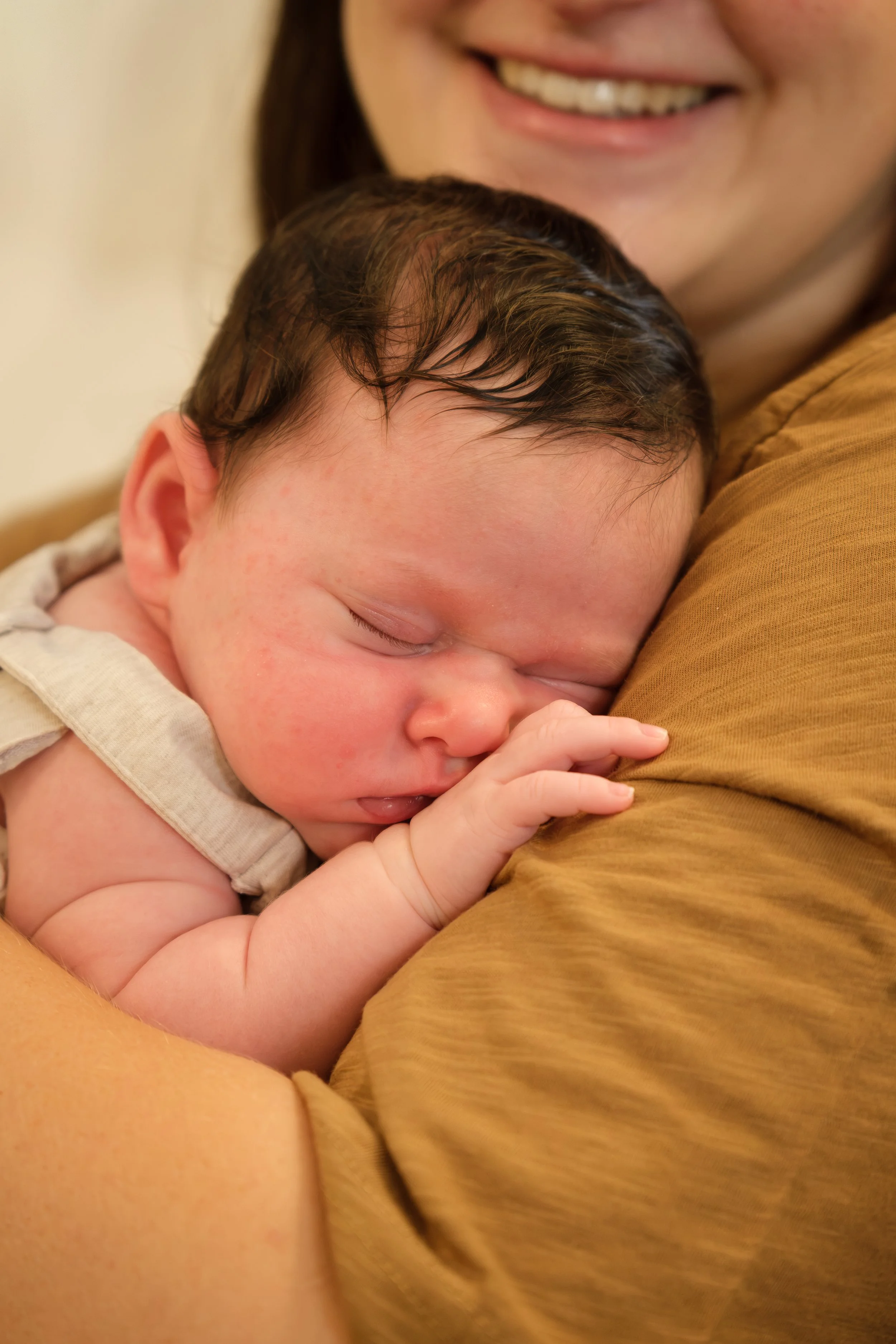 A baby sleeping peacefully on an adult's shoulder with a smile in the background.