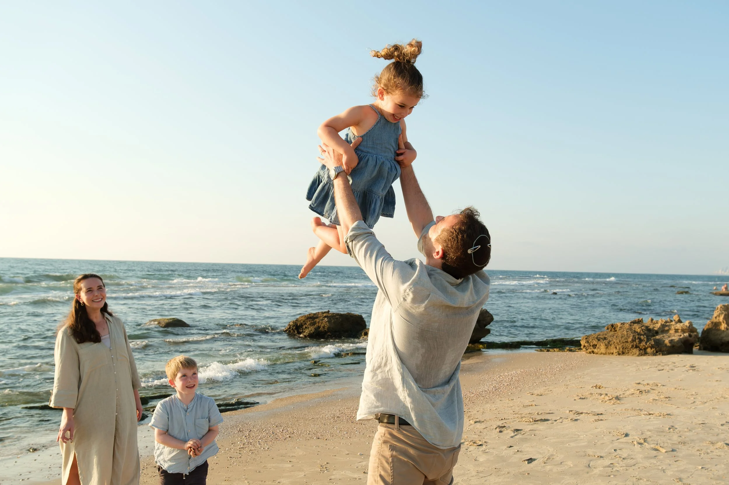A group of people at the beach, with a man lifting a young girl into the air, and two children and a woman smiling nearby.