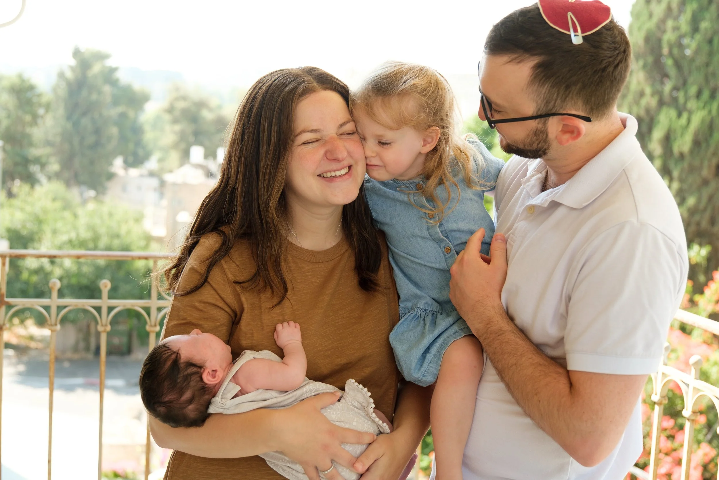 A family of four, including a mother, father, a young girl, and a newborn, enjoying a joyful moment outdoors on a bright day with trees and a white fence in the background.