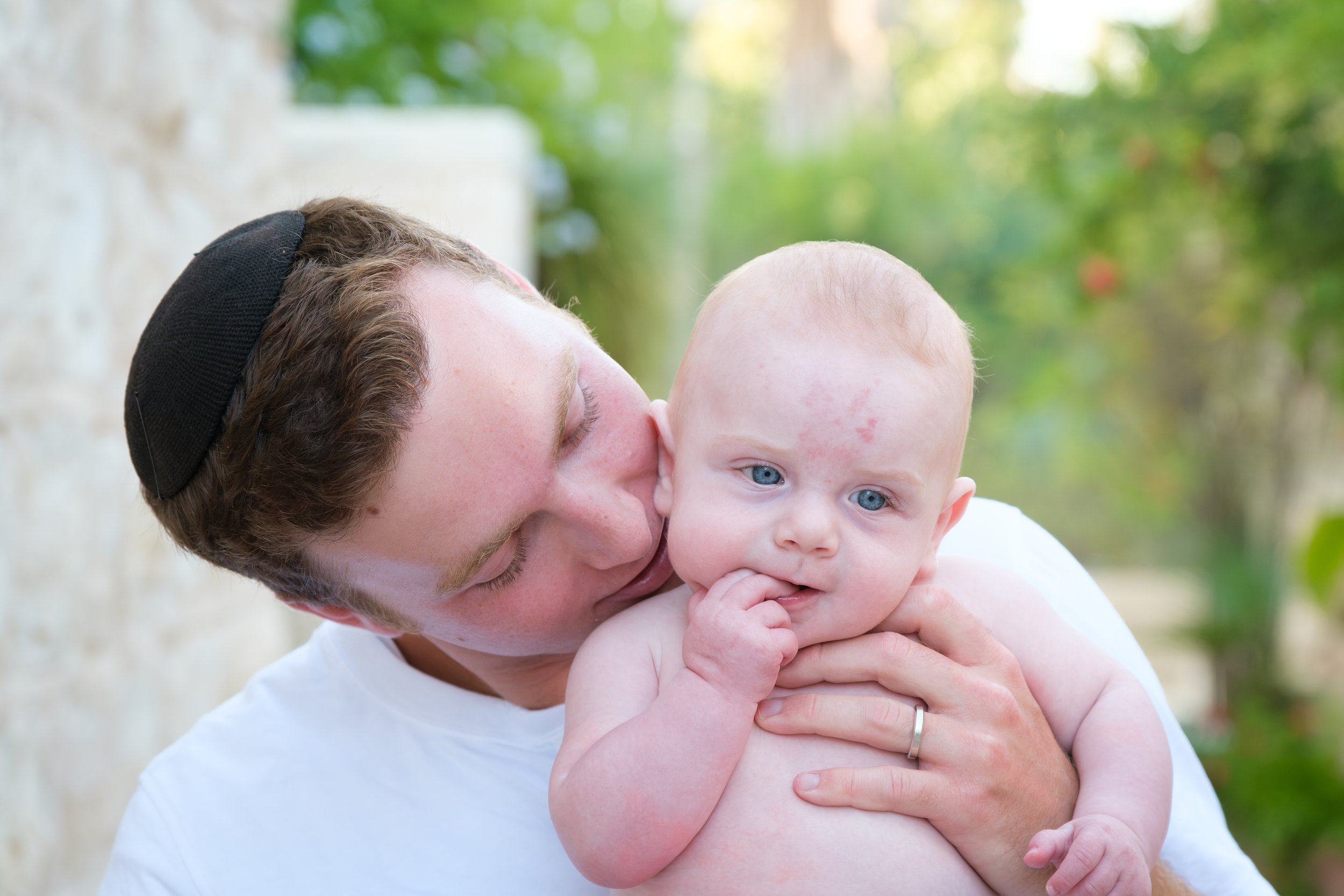A young man with curly hair, wearing a black cap, holding and kissing a baby with blue eyes and light hair outdoors with a blurred green background.