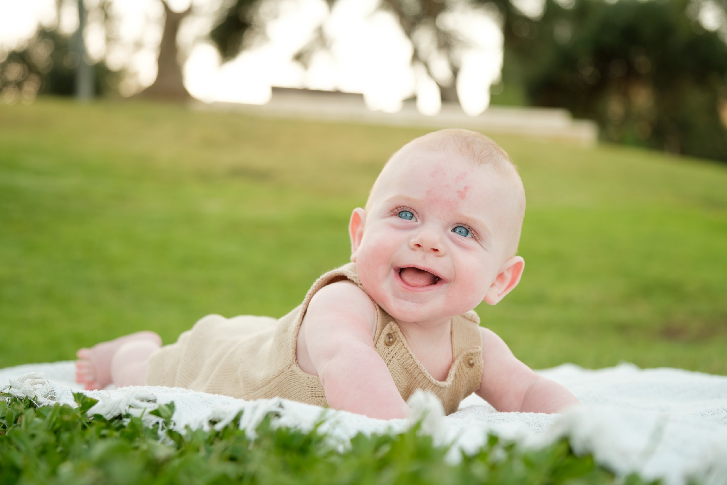 A smiling baby with blue eyes and light hair, crawling on a white blanket outdoors on green grass with trees and sunlight in the background.