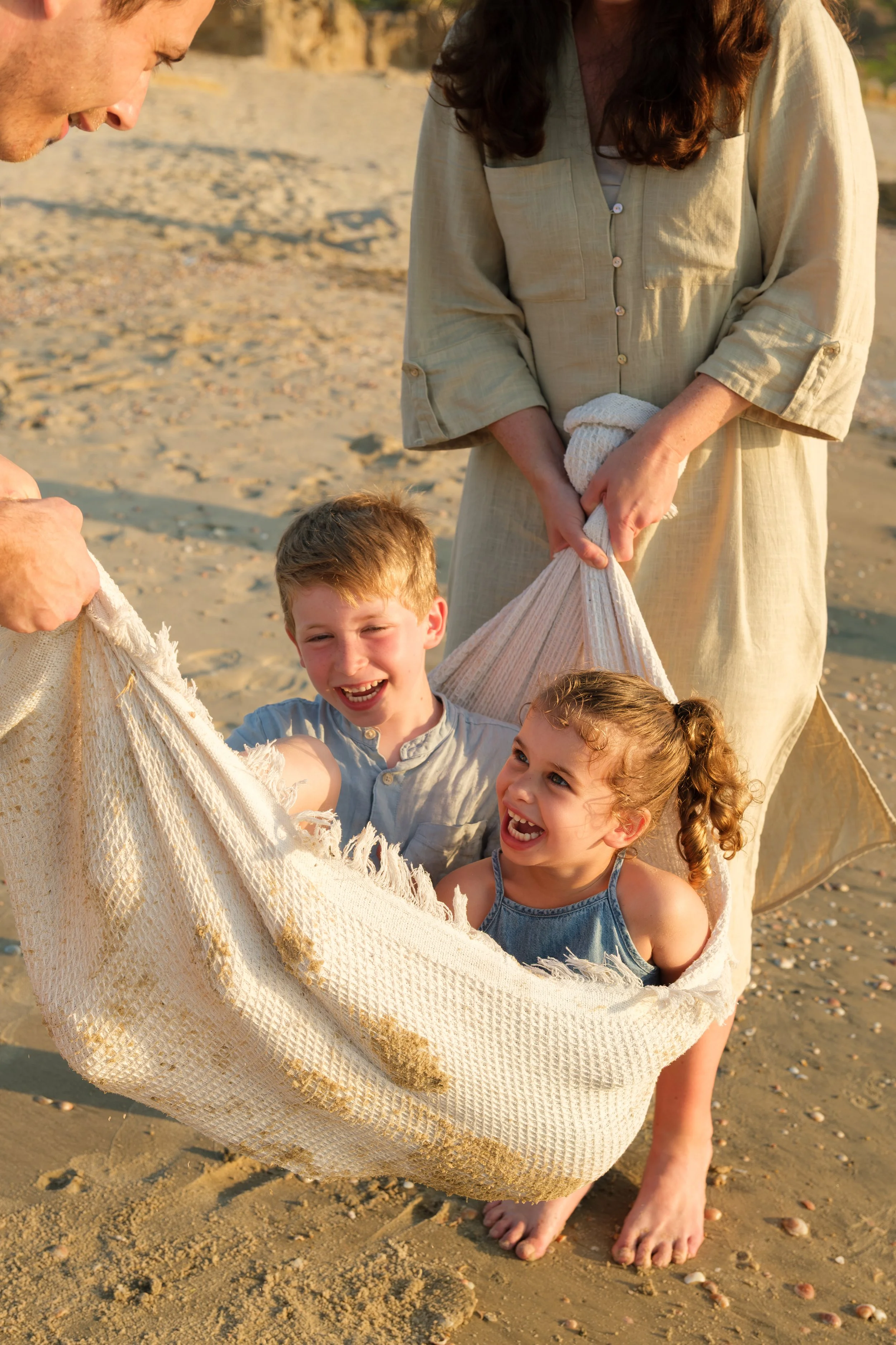 Children and adults standing on a sandy beach holding a large, woven, cream-colored blanket.