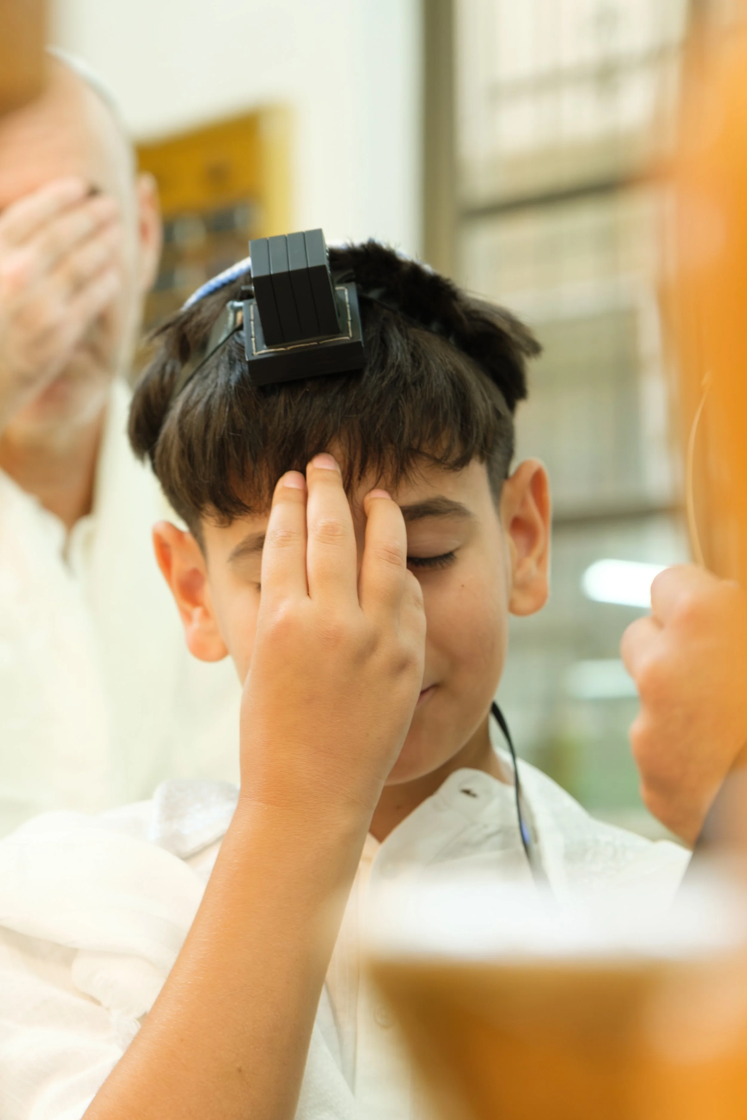 A young boy with closed eyes is in a classroom or office, with a person on the left side of the frame, possibly applying a headset or some other device to his head. The boy has dark hair and is wearing a white shirt. The background shows a window wit