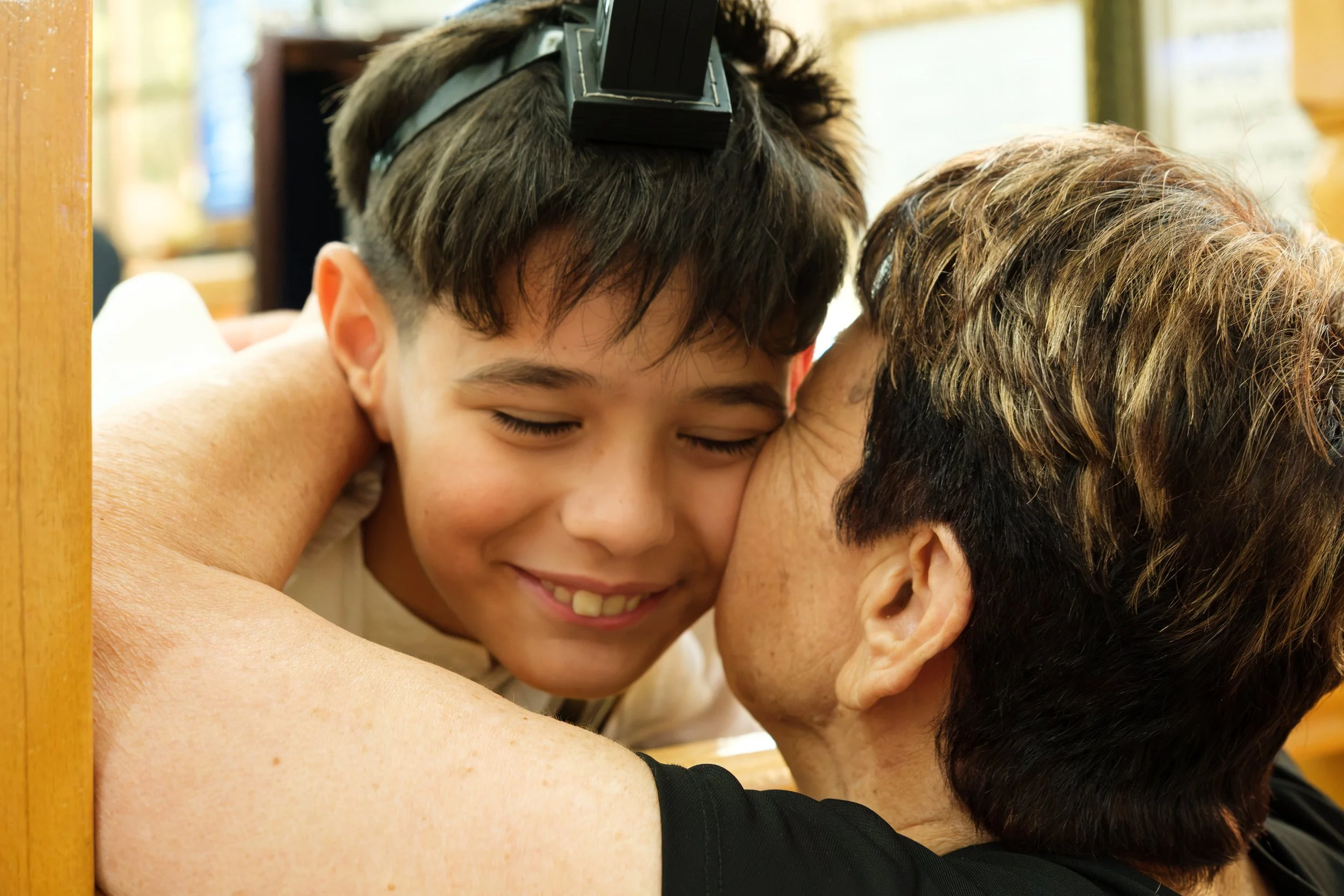 A young boy hugging an adult woman, both smiling with eyes closed, in an indoor setting with sunlight.