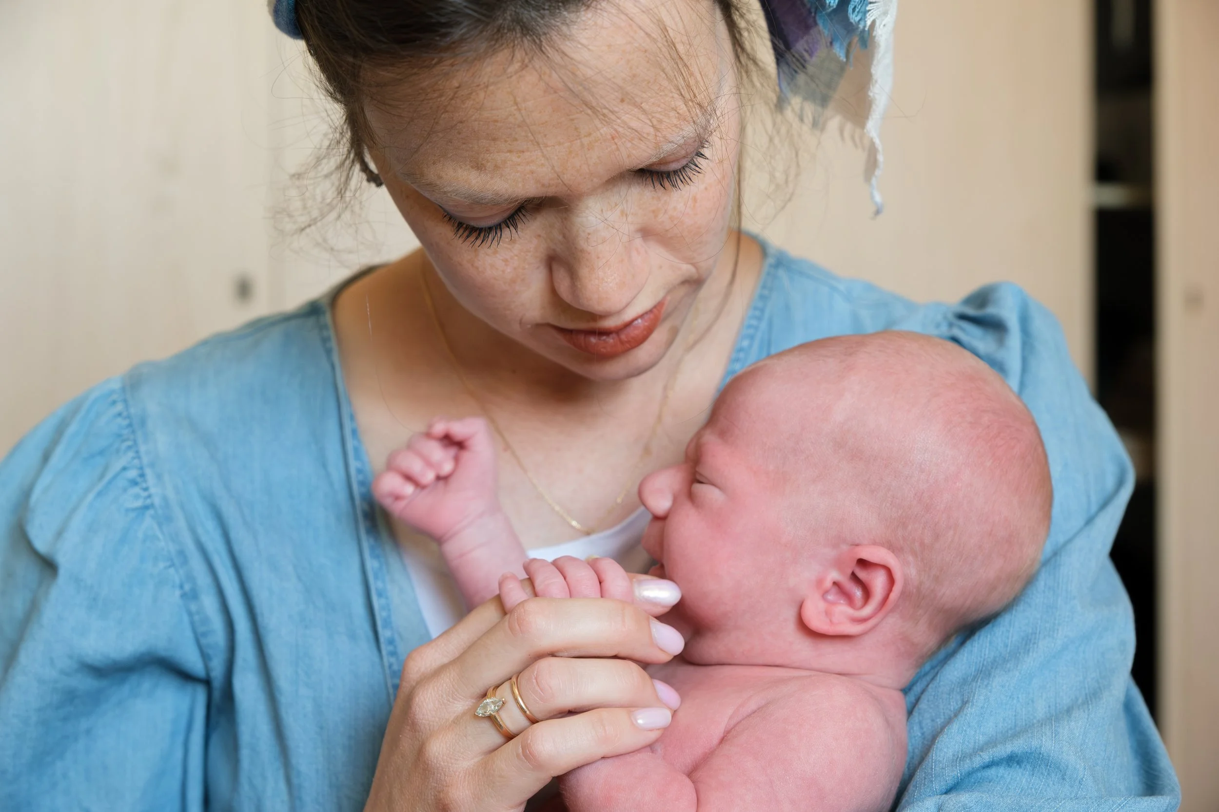 A woman with freckles and long eyelashes holds a newborn baby close to her chest, touching the baby's head affectionately.