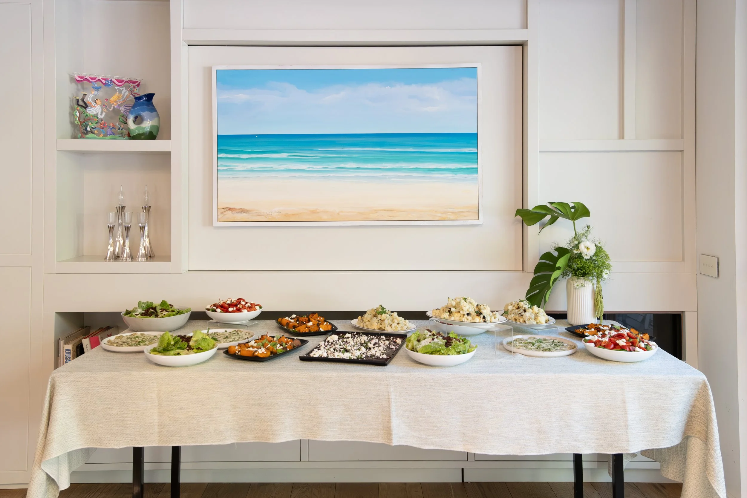 A buffet table with various salads and dishes in a dining room, decorated with a large beach scene painting on wall, shelving with dishes and decorative items, and a potted plant.