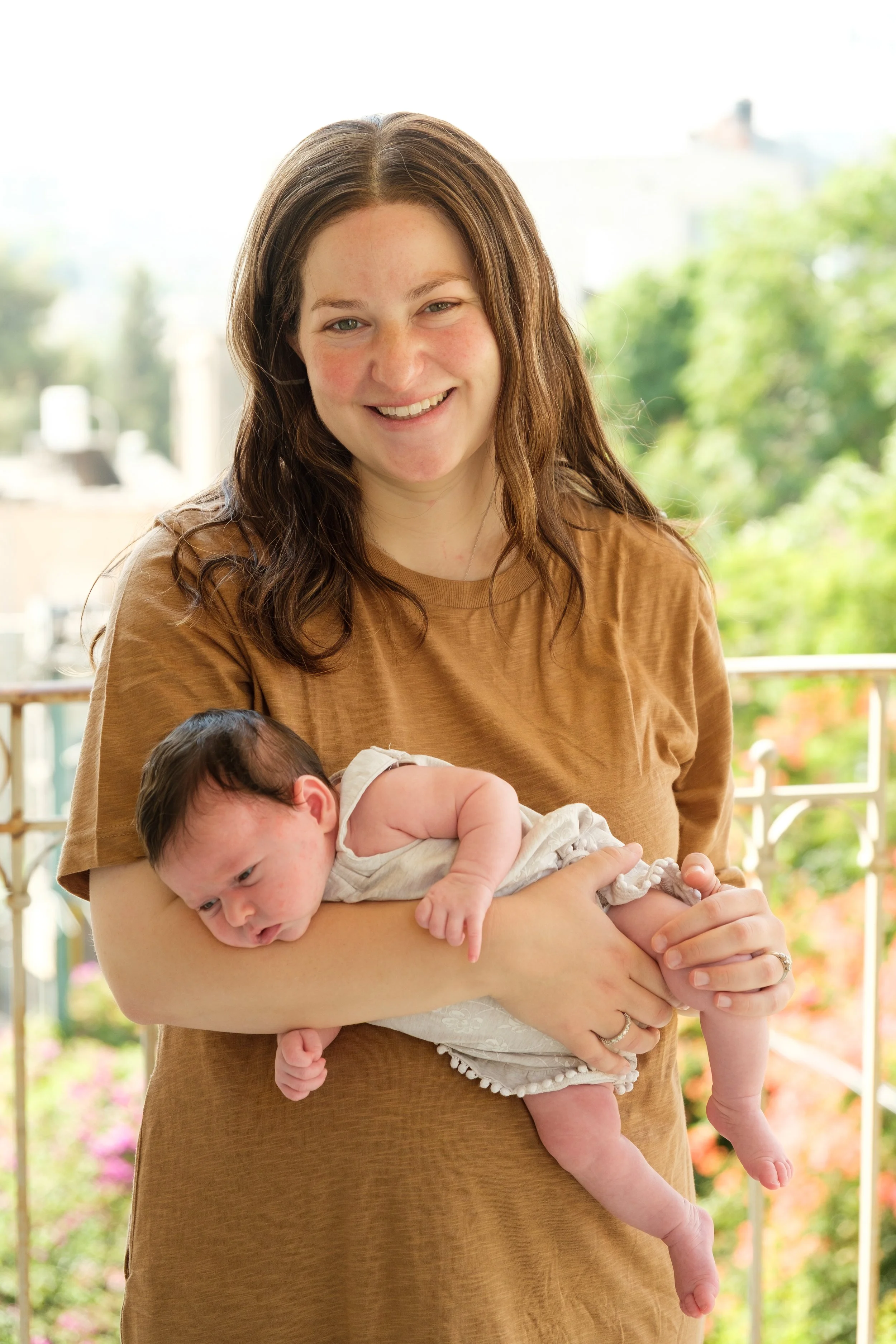 A woman smiling while holding a newborn baby outdoors on a sunny day with green trees and a white railing in the background.