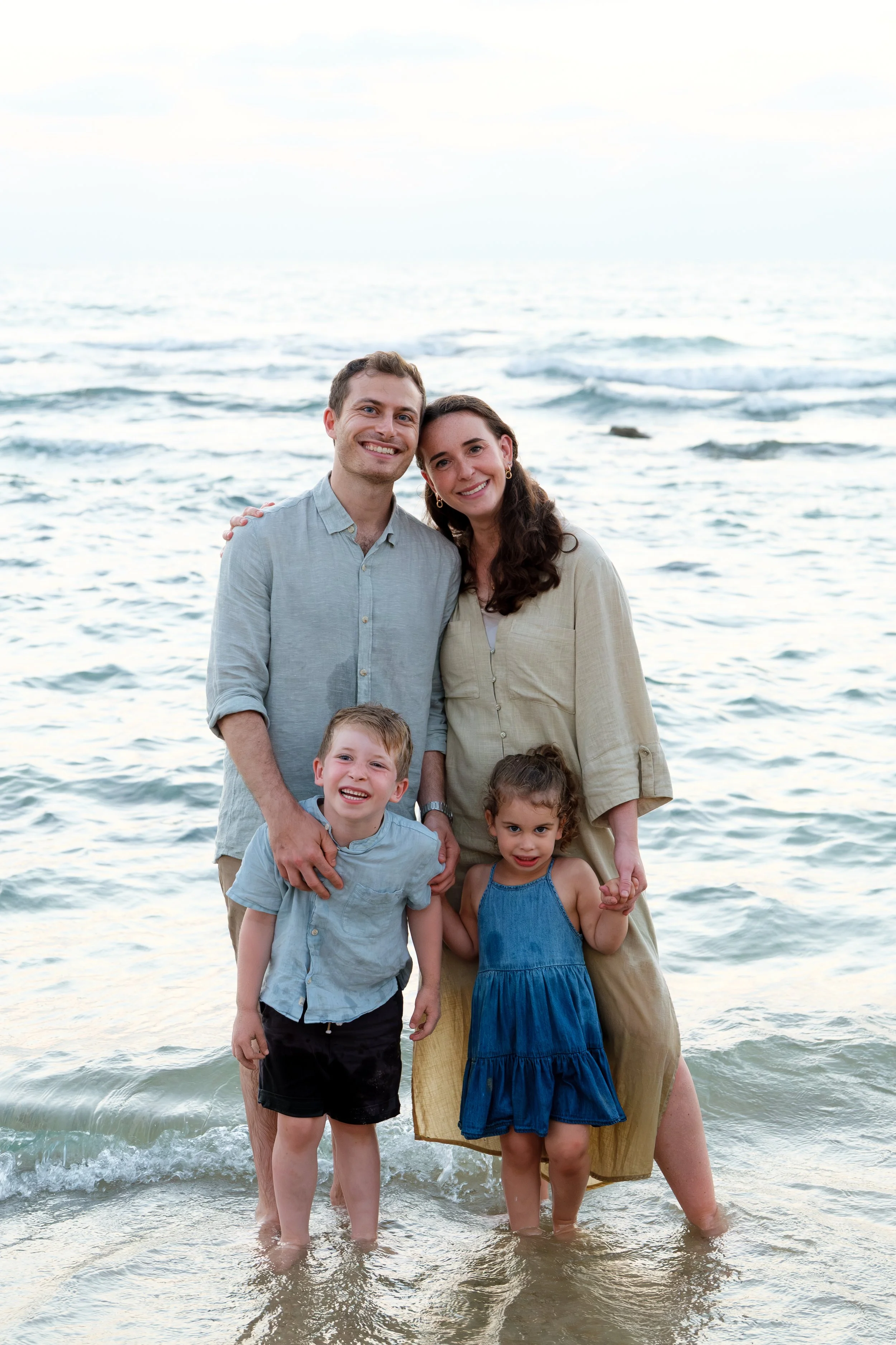 Family of five standing in the ocean near the shore, smiling at the camera. Two adults and three children, with waves behind them and a cloudy sky.