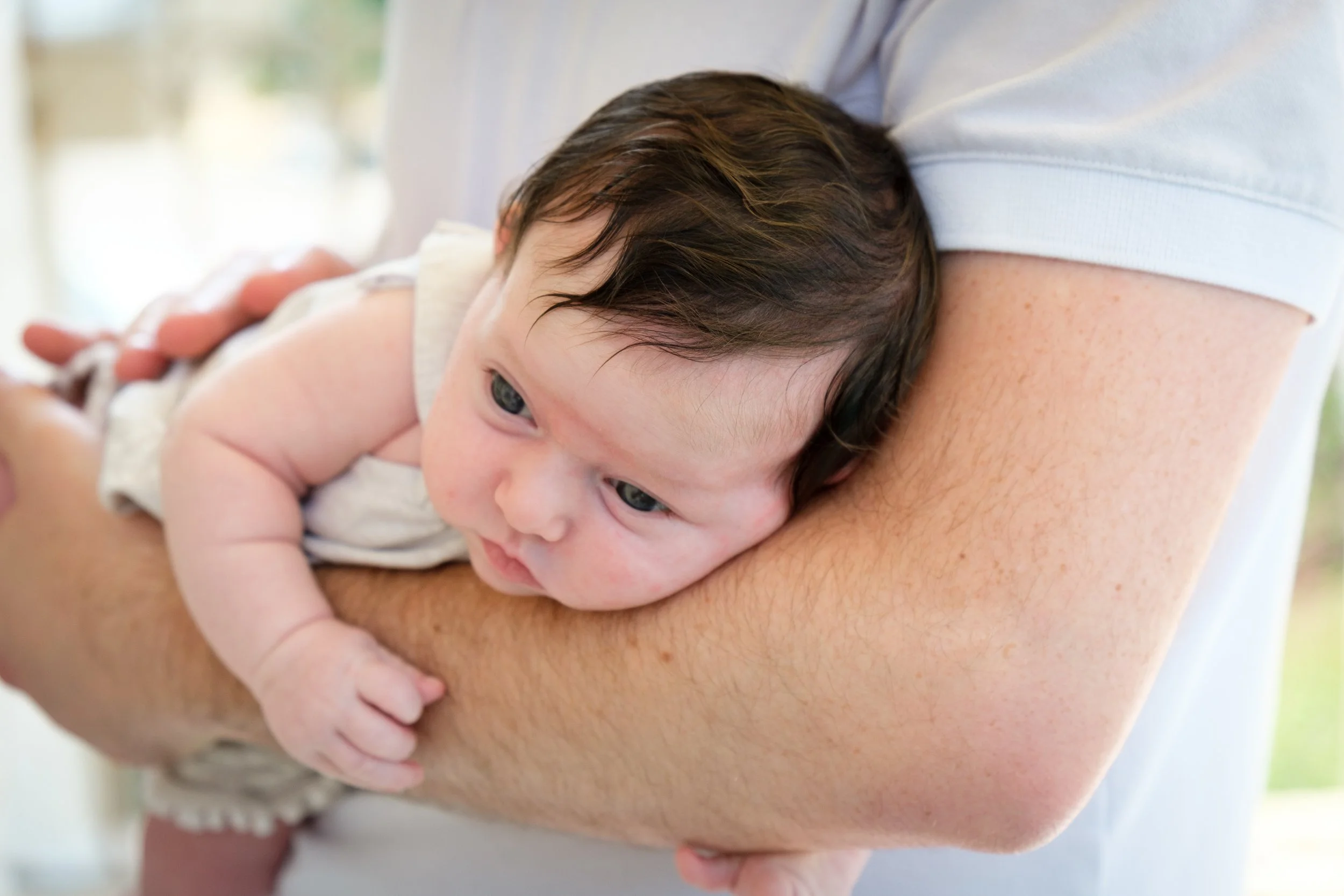 A young baby with brown hair and blue eyes resting on an adult's arm, looking to the side.