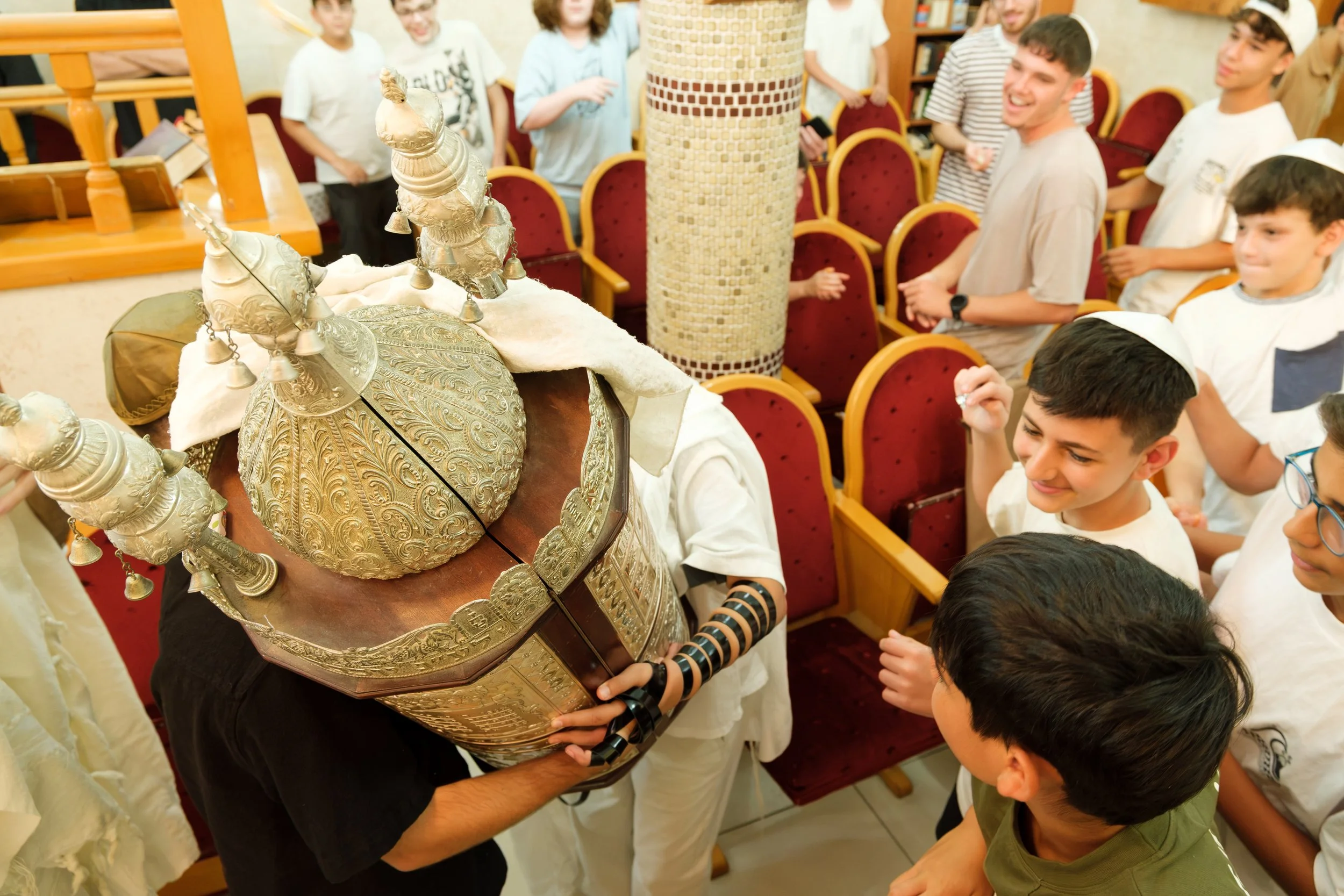 Celebration of a Jewish event with children and adults gathered around a person holding a decorative Torah scroll.