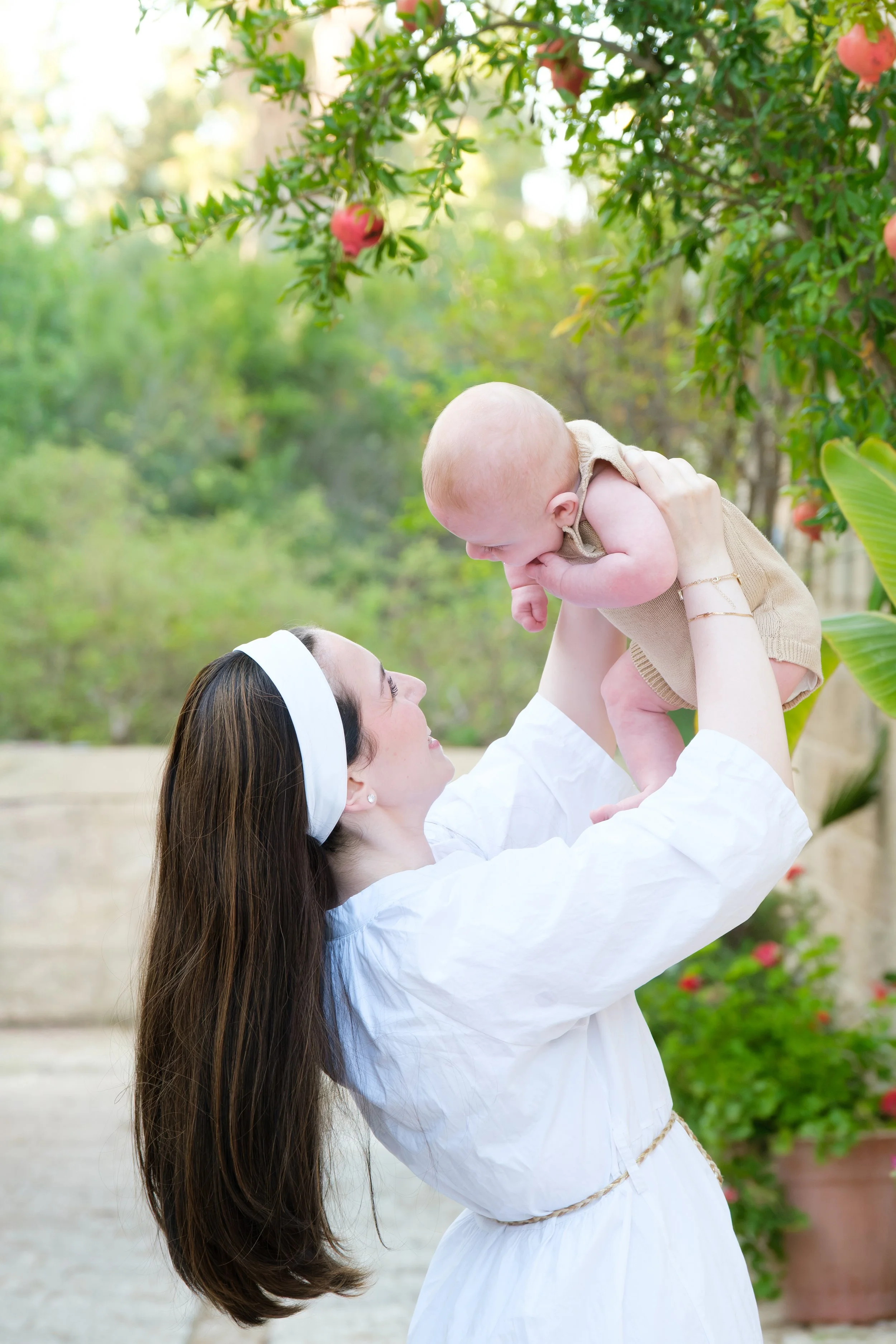 A woman with long brown hair wearing a white dress and headband is lifting a baby girl in a tan romper towards her in a garden with greenery and pink flowers.
