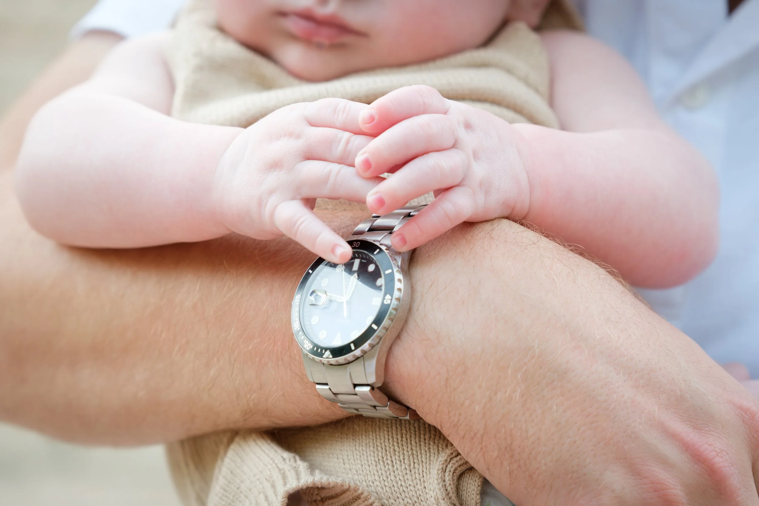 A young child with light skin and blonde hair wearing a beige vest and white shirt, holding hands with an adult and looking at a wristwatch on the adult's wrist.