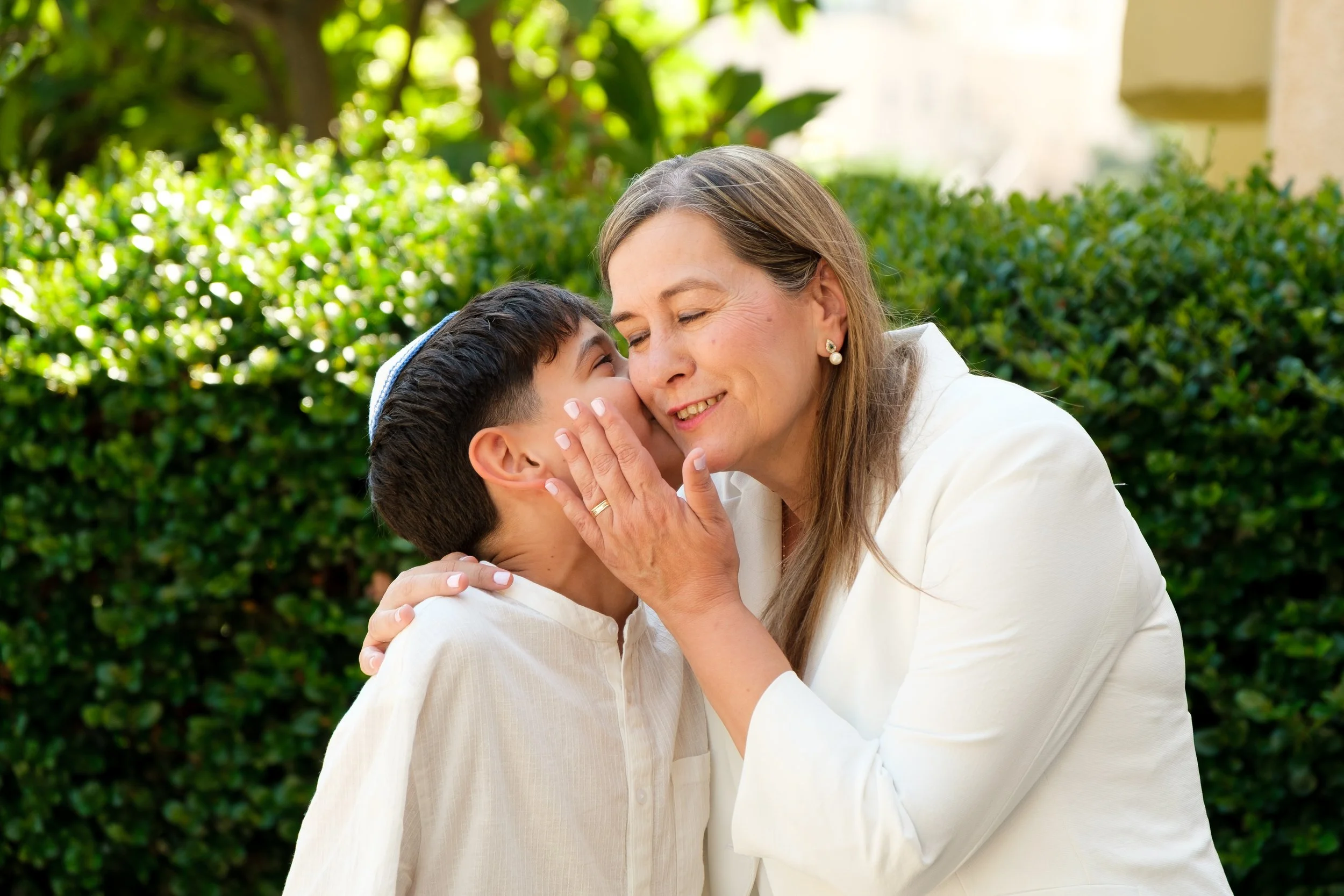 A woman hugging and kissing a young boy on the cheek outside with green bushes and trees in the background.