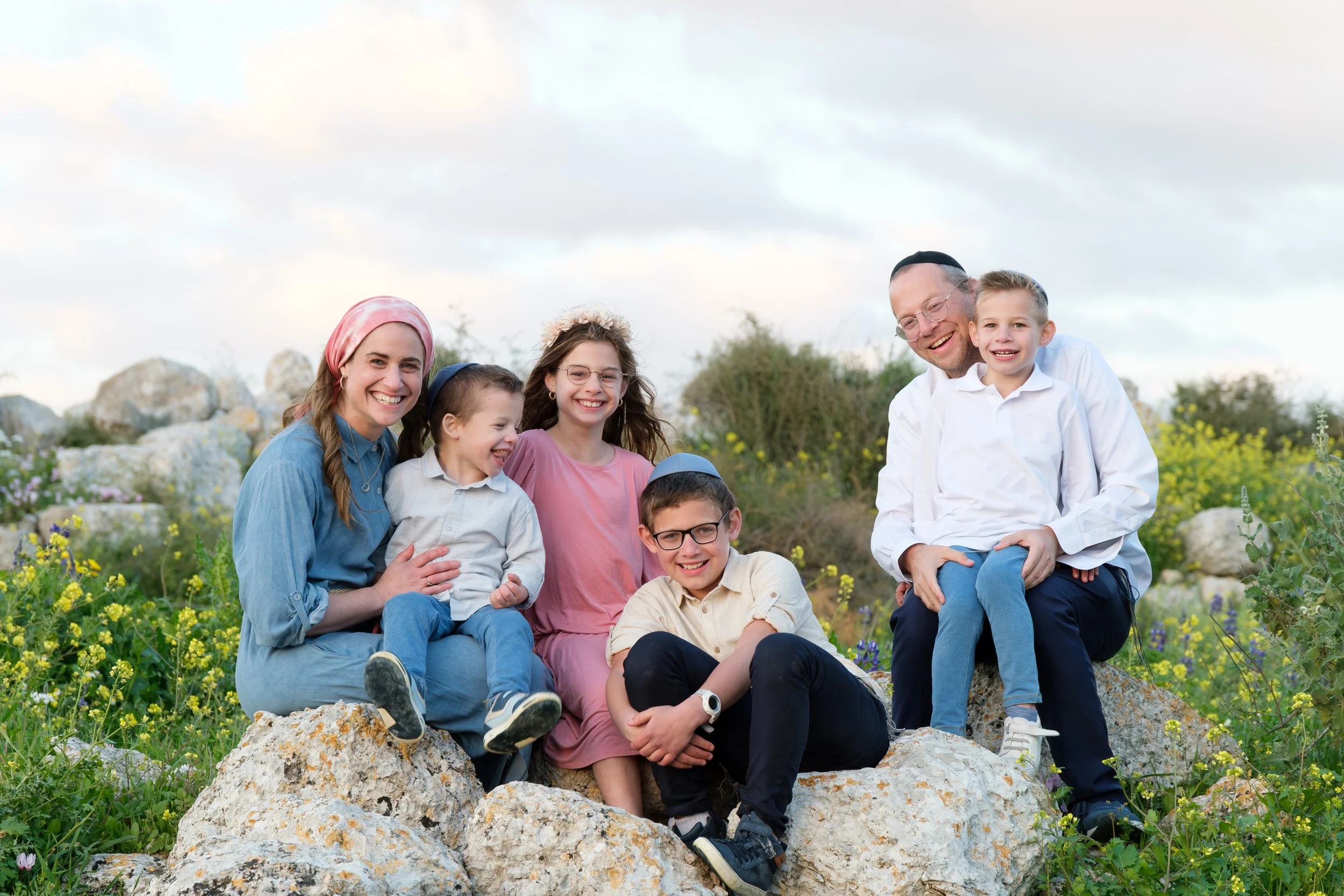 A group of seven people, including children and an adult, sitting and standing on rocks in a field of green plants and yellow flowers, smiling at the camera.
