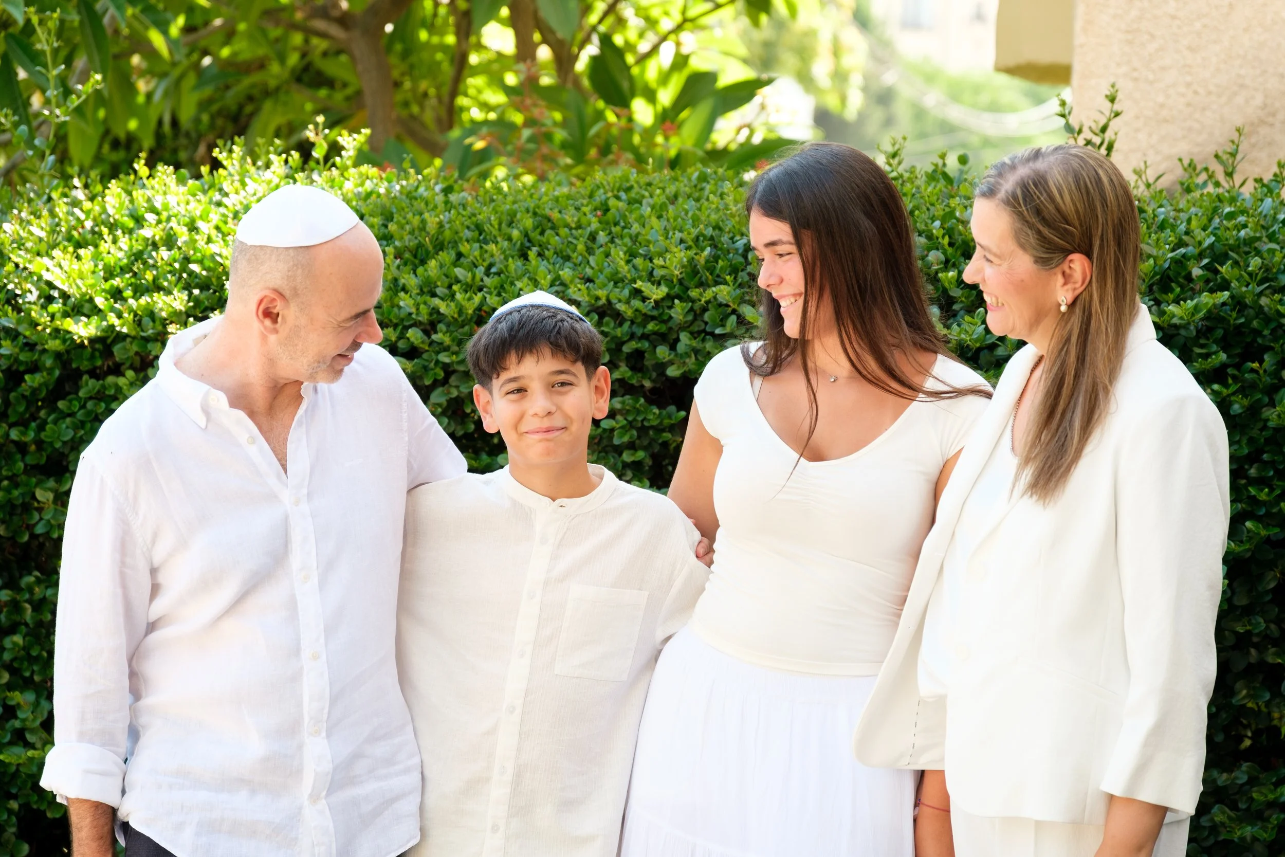 A Jewish family outdoors, including an older man with a kippah, a young boy, a woman in white, and another woman with earrings, smiling and enjoying a sunny day near greenery.