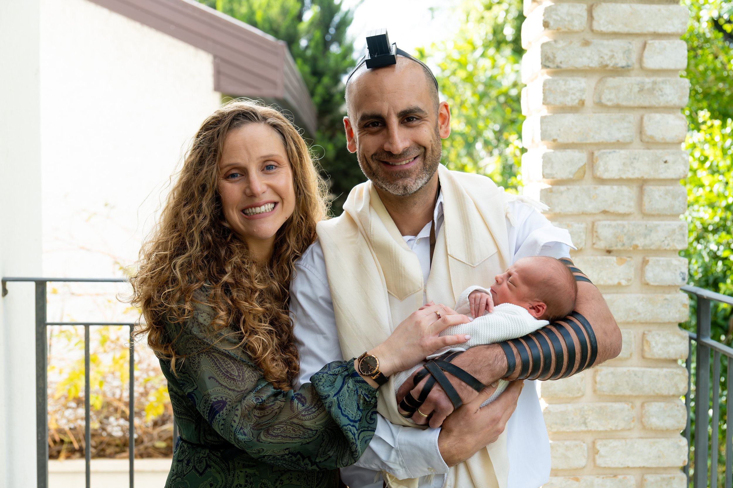 A man with a bald head and a beard holding a newborn baby, standing outdoors next to a woman with long curly hair. The man has a small solar panel on his head, and they are smiling at the camera. The background features trees and a brick column.
