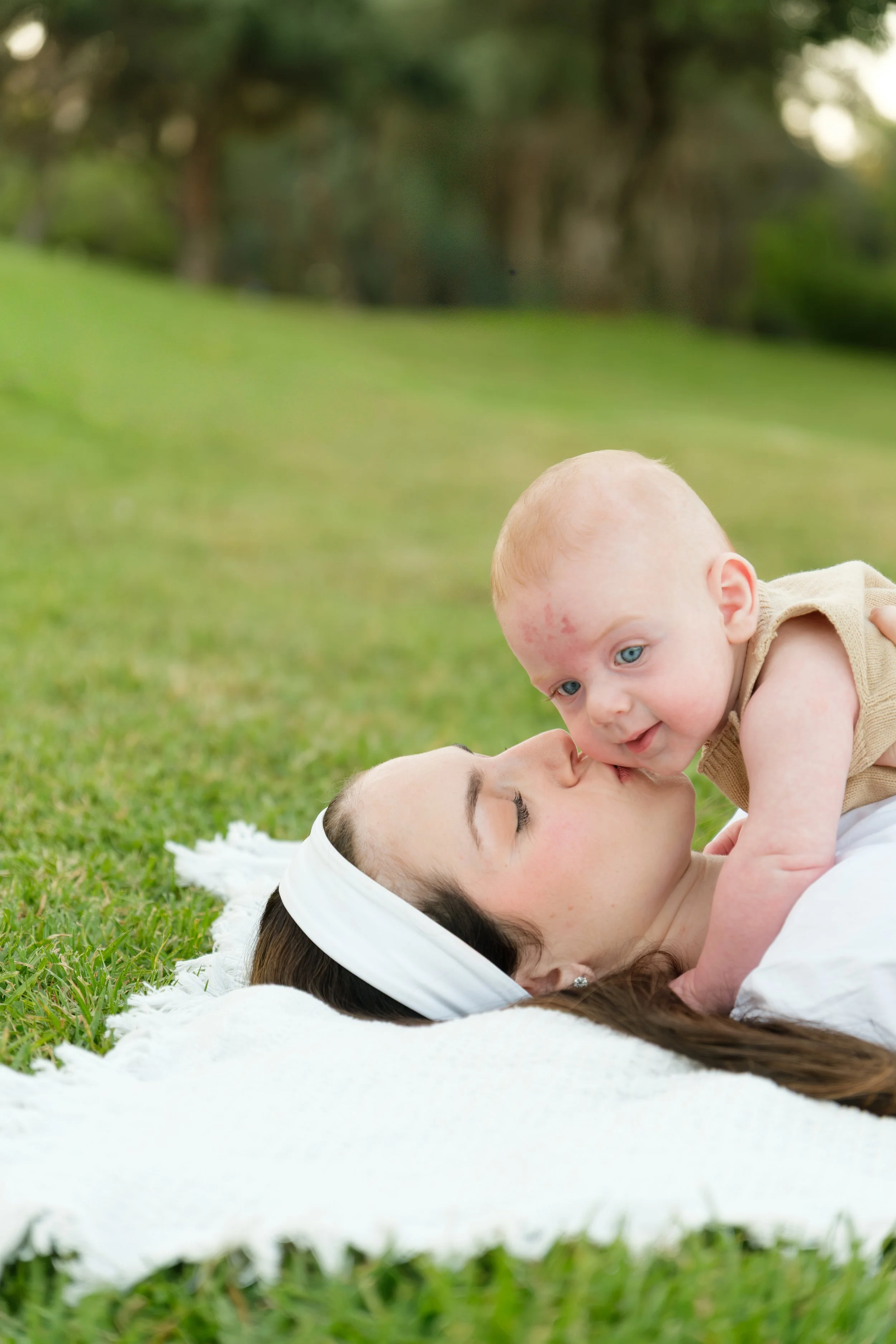 A woman lying on the grass with a white headband, kissing a baby boy on the nose during daytime outdoor setting, blurred green trees in the background.
