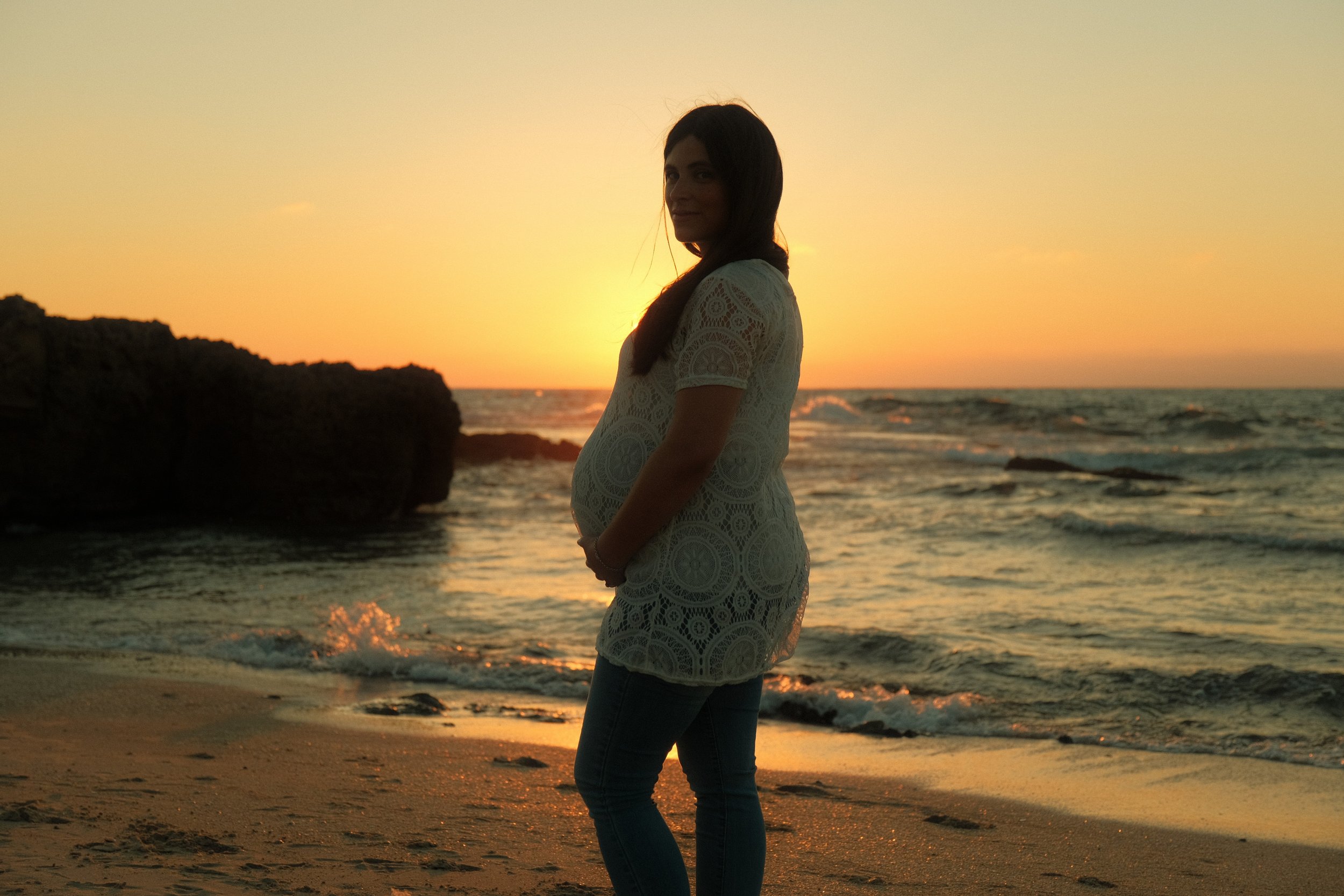 A woman in a lace dress standing on the beach during sunset, with her profile visible and the ocean in the background.