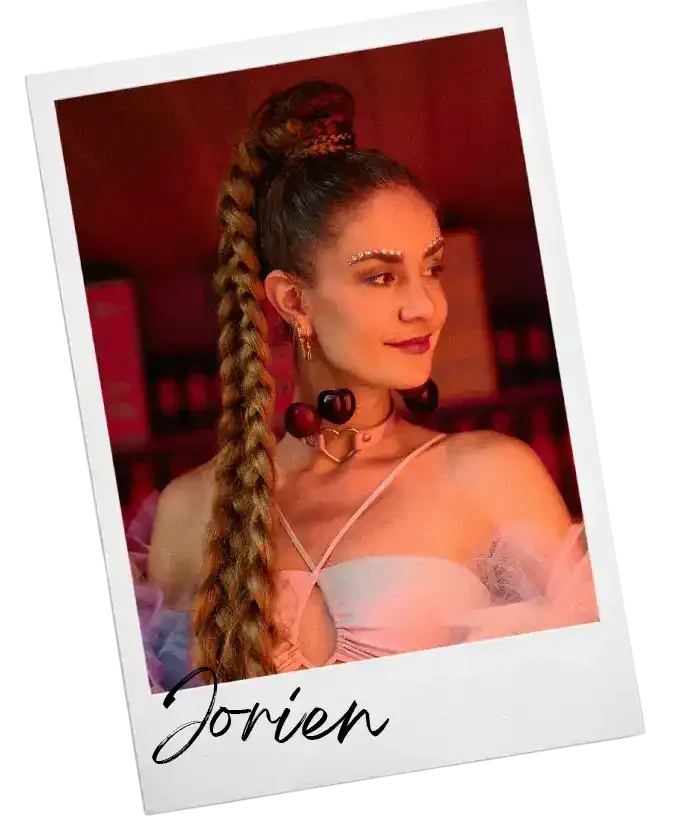 Een vrouw met lang, gekruld haar in een hoge paardenstaak, versierd met bloemen, draagt sieraden en een roze top. Ze staat in een kleurrijke, mogelijk feestelijke setting.