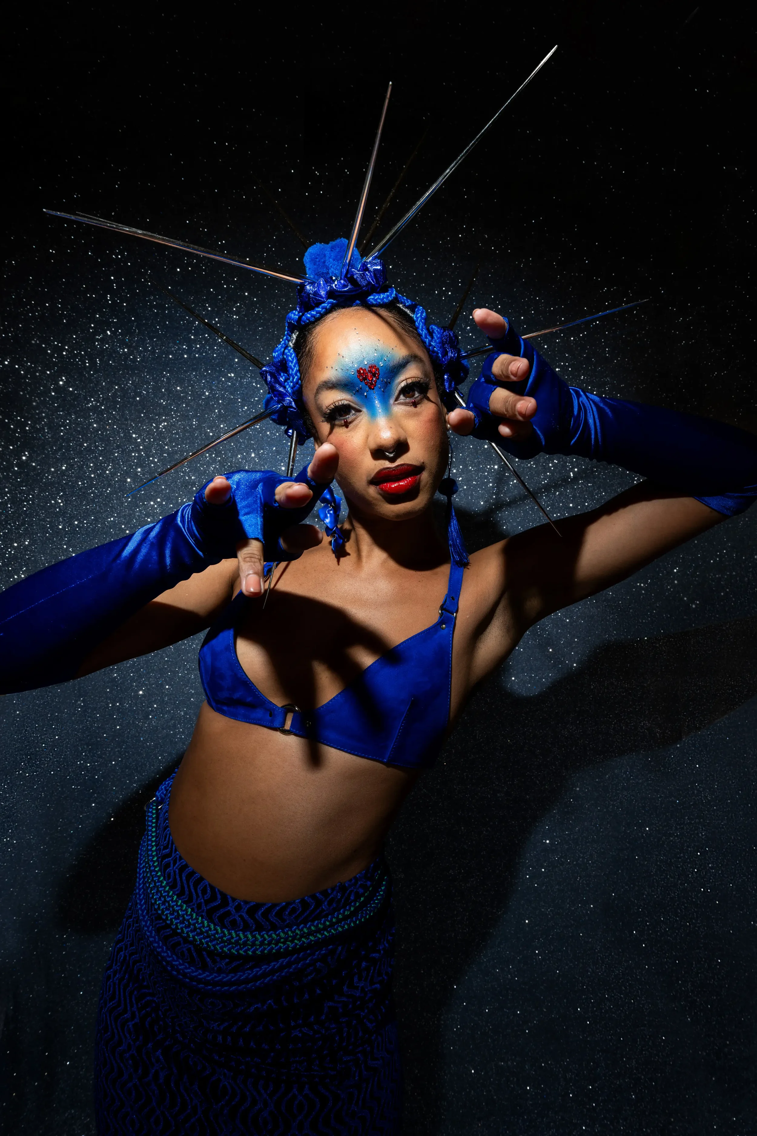 Een vrouw in een opvallende blauwe outfit met creatief stijladvies, haar gezicht versierd met blauwe make-up en een hartvormig patroon op haar voorhoofd, poseert tegen een sterrengekleurde achtergrond.
