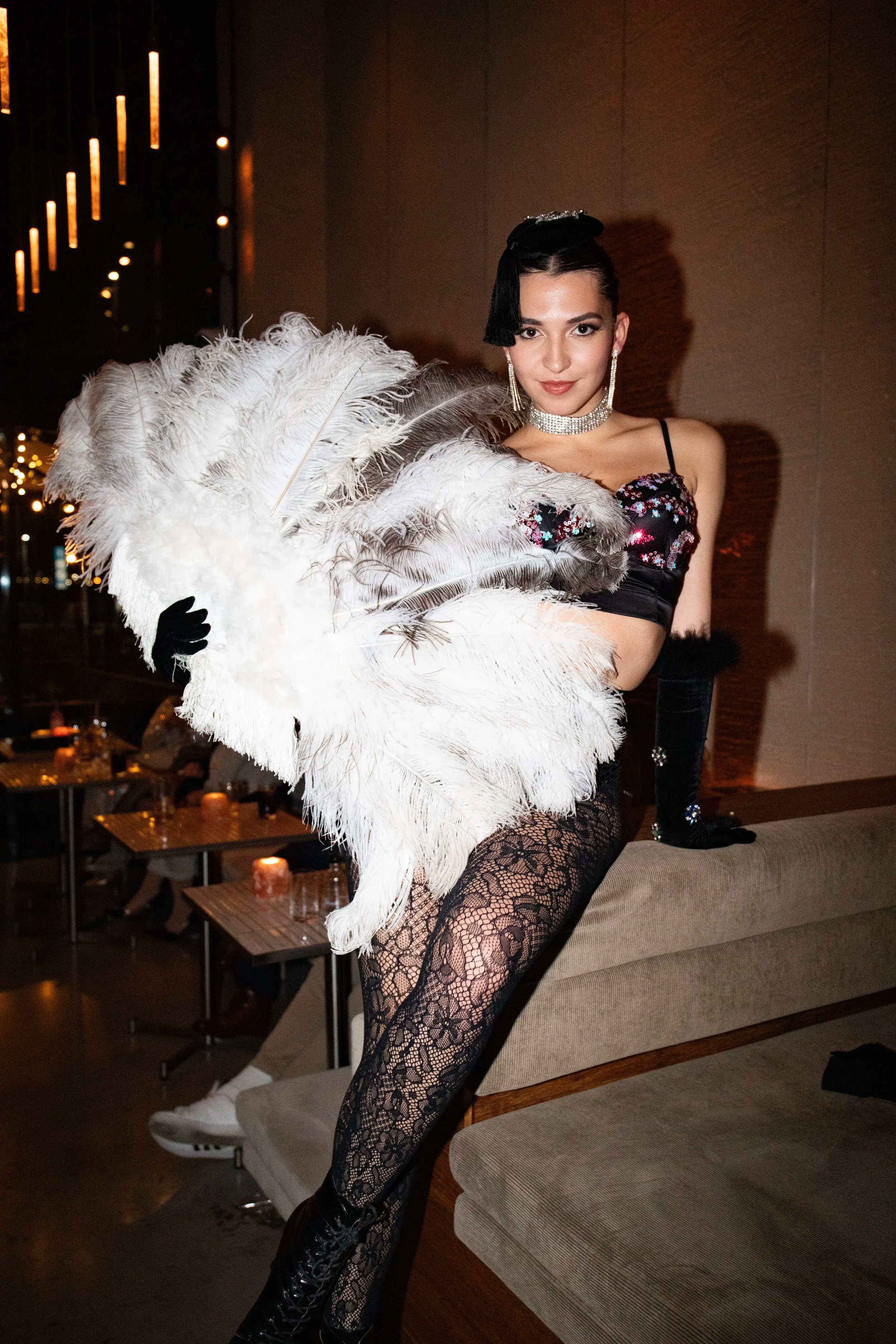 Vrouw in een feestoutfit met zwarte lace panty's, een krokodillenleren laarzen, parelketting, oorbellen en een haaraccessoire, die post met een grote veerfans, in een dimmige, feestelijke setting.